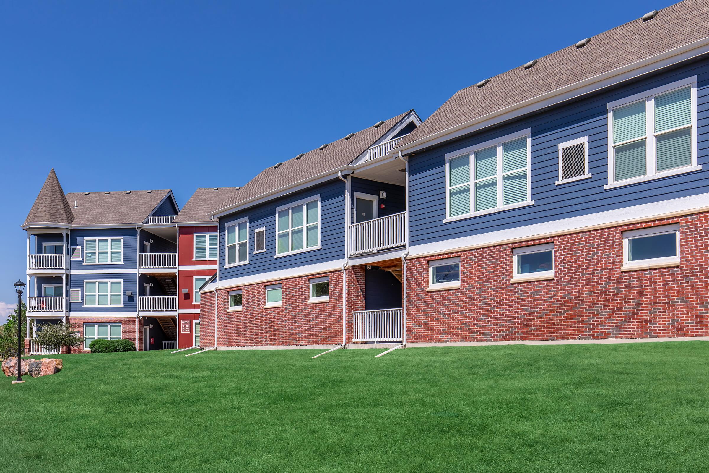 Row of multi-unit residential buildings featuring various architectural styles. The foreground shows a well-maintained grassy area, while the buildings feature brick and siding exteriors. Blue sky with minimal clouds enhances the bright, sunny atmosphere.