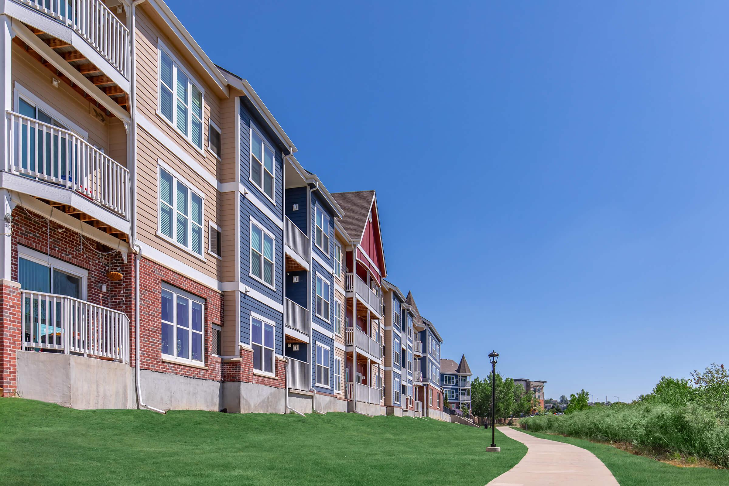 A row of multi-story apartment buildings in various colors, including blue, beige, and red. The buildings feature balconies and large windows. In the foreground, there is a paved walkway bordered by green grass and a lamp post, set against a clear blue sky.