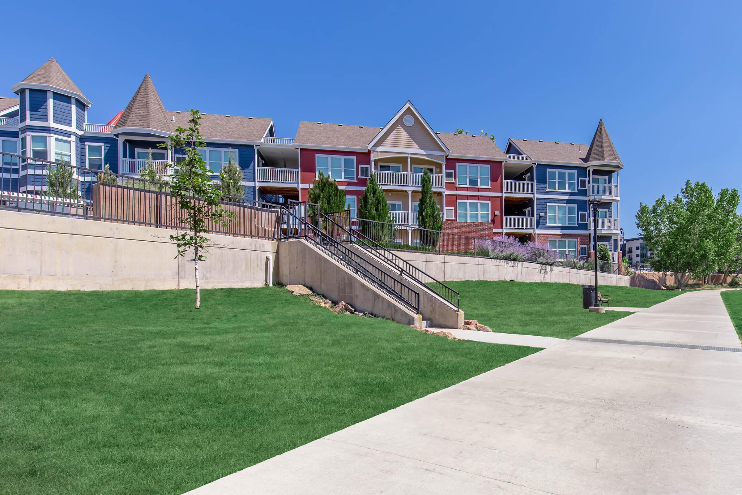 A modern residential building featuring colorful exteriors and multiple balconies, situated on a landscaped green lawn. A pathway runs alongside the property, leading up to the entrance, with a ramp for accessibility. Clear blue skies enhance the overall pleasant atmosphere.