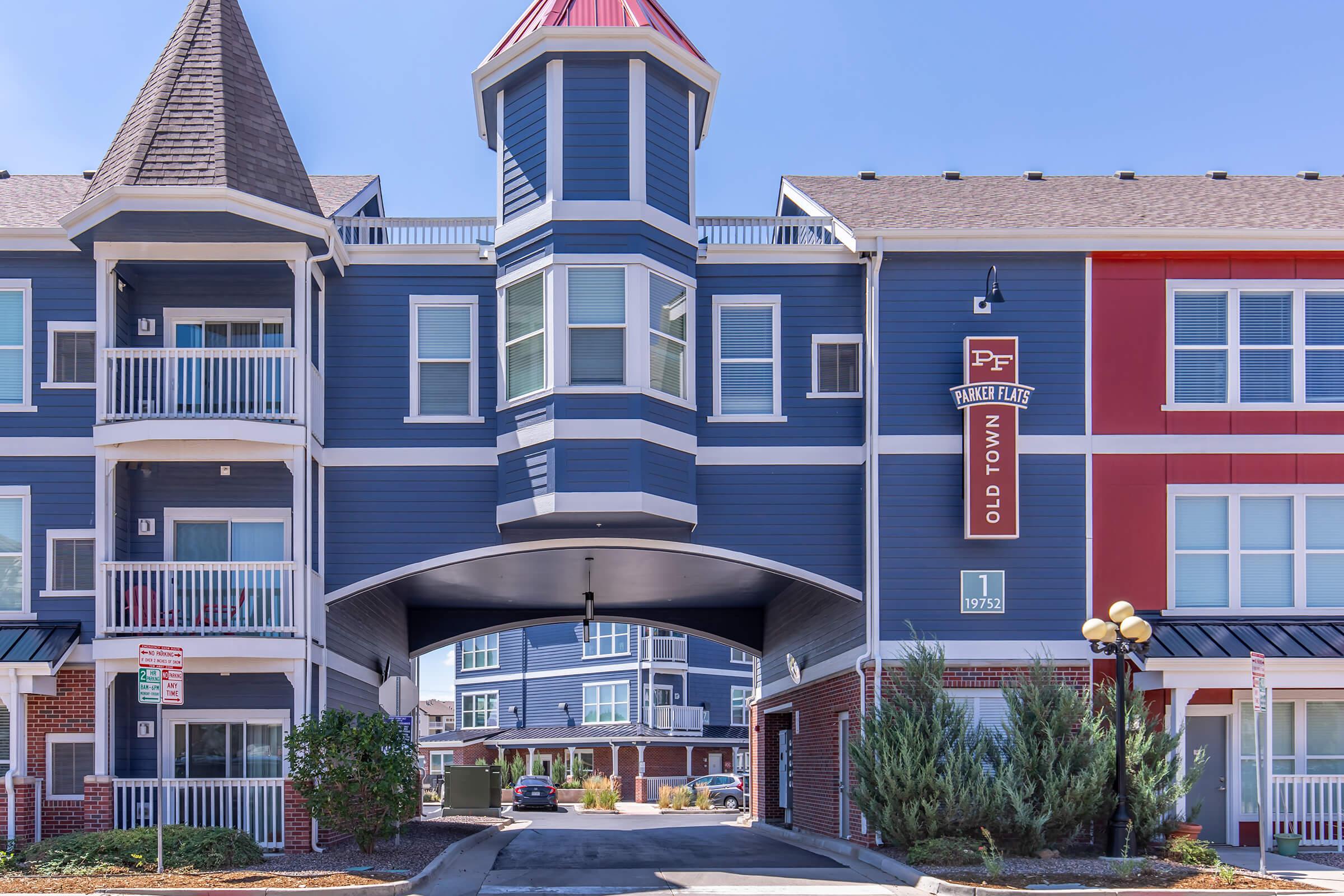 Modern multi-story apartment building featuring blue and red exteriors, with a prominent tower structure in the center. The area underneath the tower serves as an entrance, with balconies visible on either side. Lush greenery and parking spaces are seen in the background. Clear blue sky above.