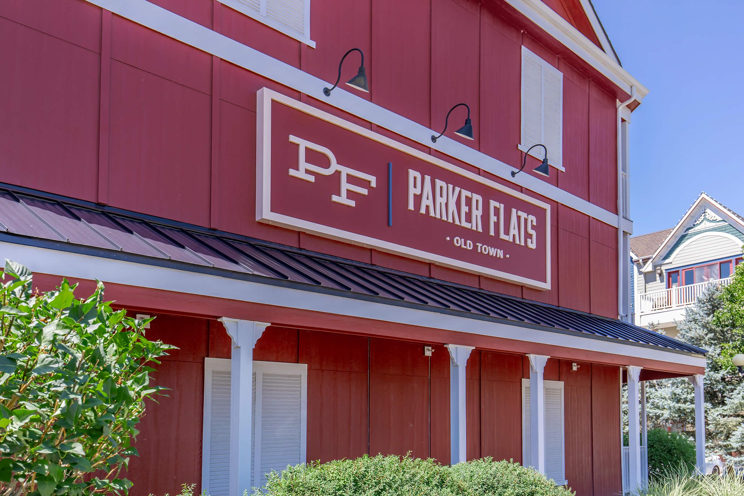 A close-up view of a red building featuring the sign "PARKER FLATS" on a white background. The sign includes the letters "PF" in a stylized font. The building has white trim and several outdoor light fixtures, surrounded by greenery, indicating a well-maintained exterior in a pleasant neighborhood setting.