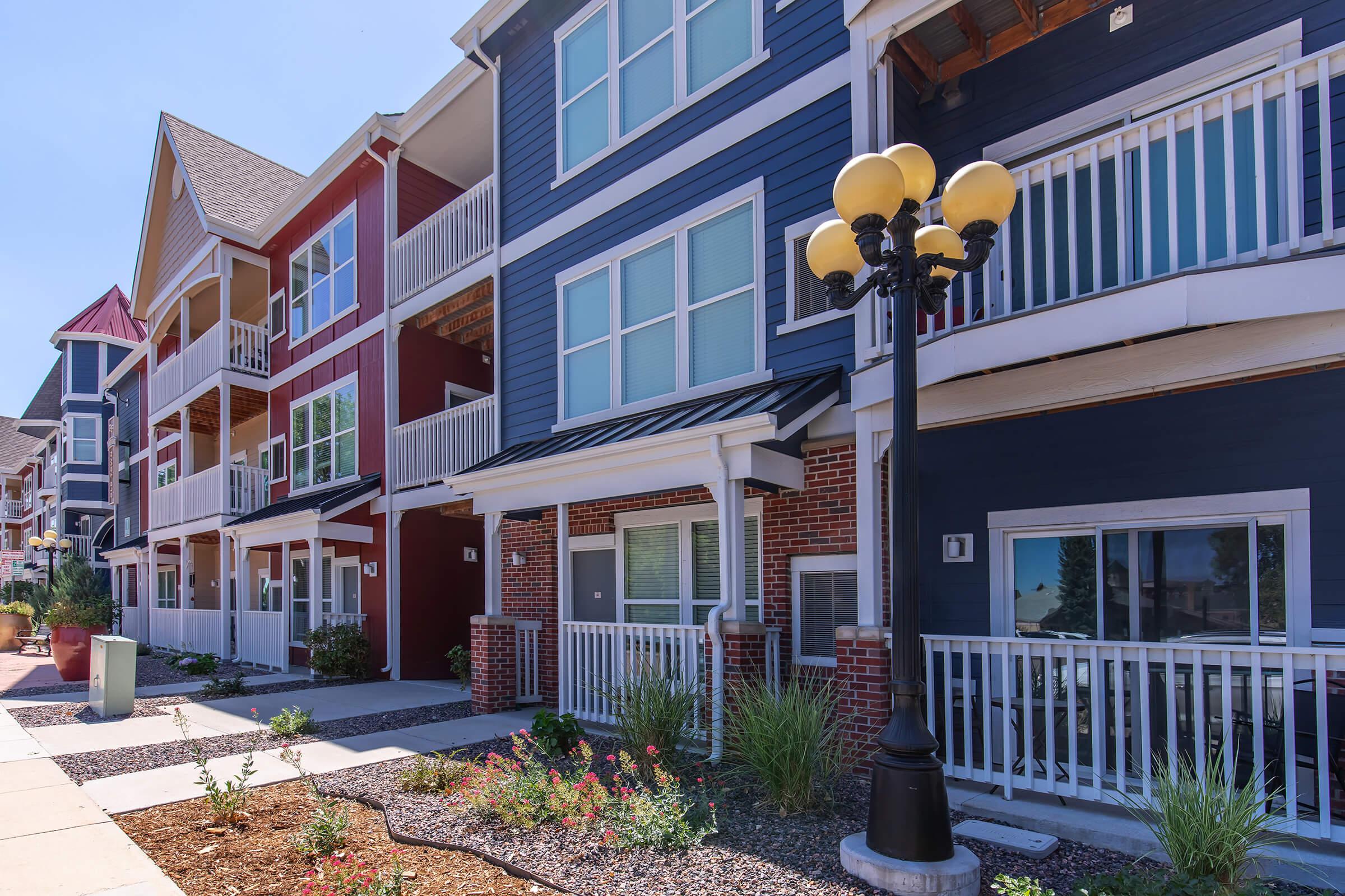 A row of modern, colorful apartment buildings featuring balconies and large windows, surrounded by neatly landscaped plants and pathways. A decorative street lamp stands prominently along the walkway. The sky is clear and blue, enhancing the vibrant appearance of the buildings.