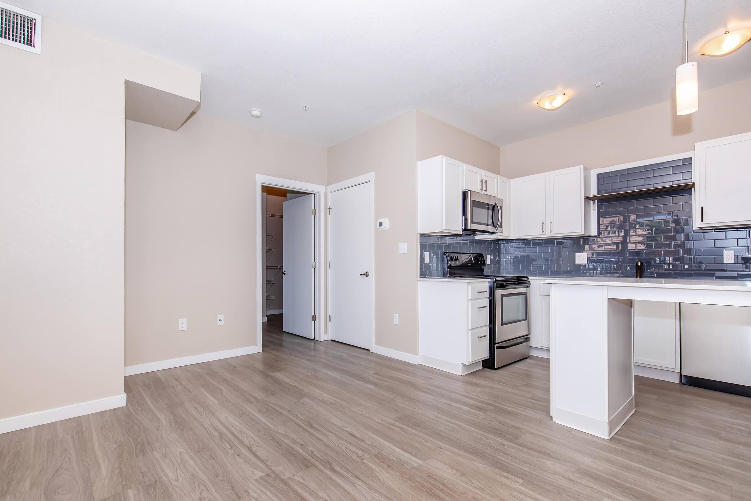 A view of a modern, open-concept kitchen and living area. The kitchen features white cabinets, a stainless steel stove, microwave, and a tiled backsplash. Light-colored walls and flooring enhance the spacious feel, with a doorway leading to another room in the background. Soft lighting fixtures brighten the space.