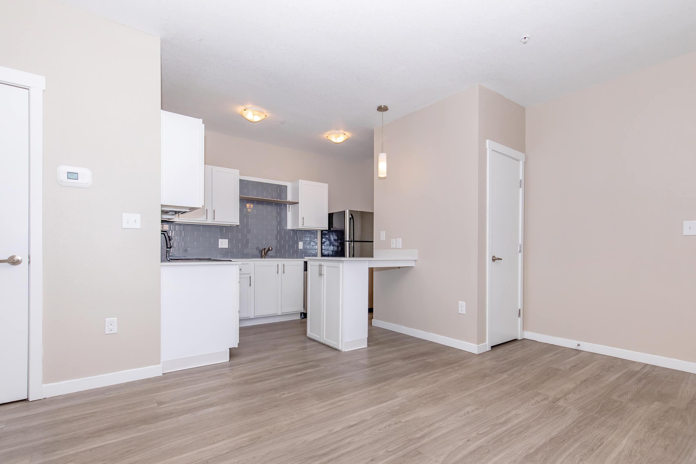 A modern, open-concept kitchen and living area featuring light-colored walls, wood-like flooring, and recessed lighting. The kitchen includes white cabinets, a grey-tiled backsplash, and a countertop that borders the living space. A door leads to another room, and a thermostat is mounted on the wall.