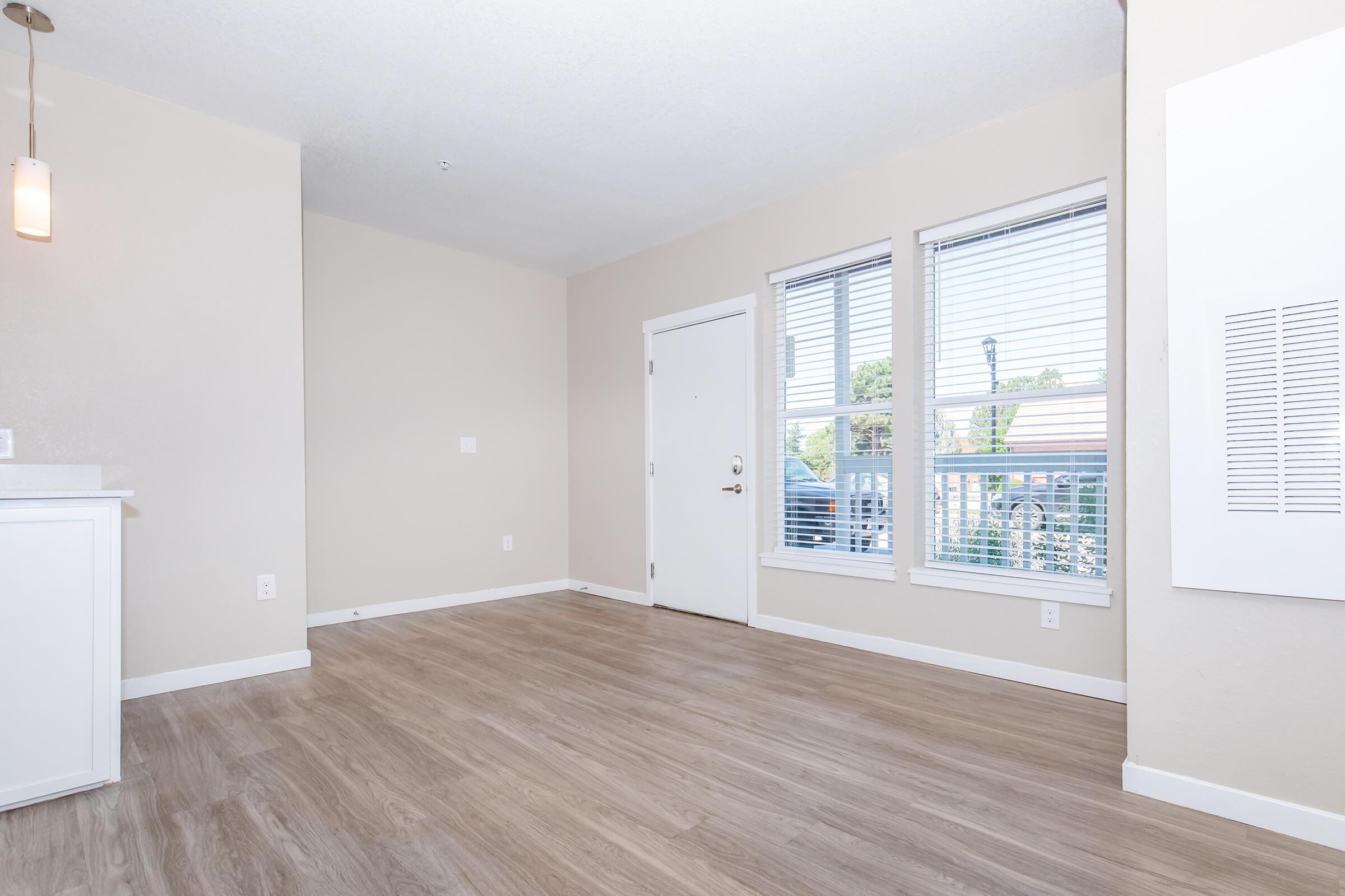 A bright, empty living room featuring light-colored walls and wood-like flooring. There are two windows with blinds allowing natural light, and a plain white wall with a door. A simple pendant light hangs from the ceiling, creating a modern and open atmosphere.