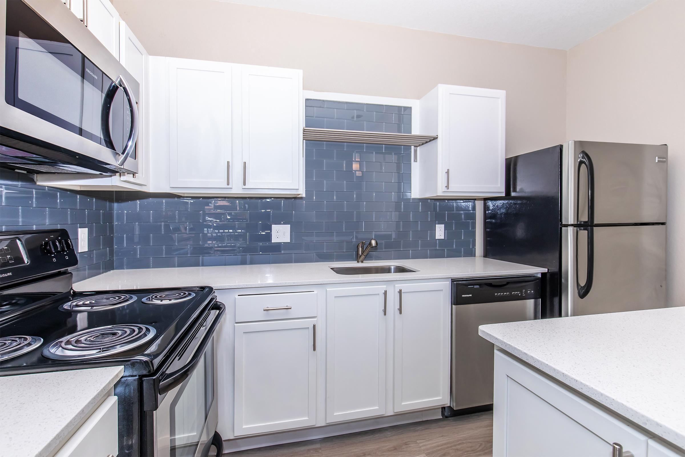 A modern kitchen featuring white cabinets, stainless steel appliances, a black refrigerator, and a dark blue tiled backsplash. The countertop is white with a subtle texture, and an oven with a smooth top is visible. The kitchen has a clean and contemporary design with ample storage and workspace.