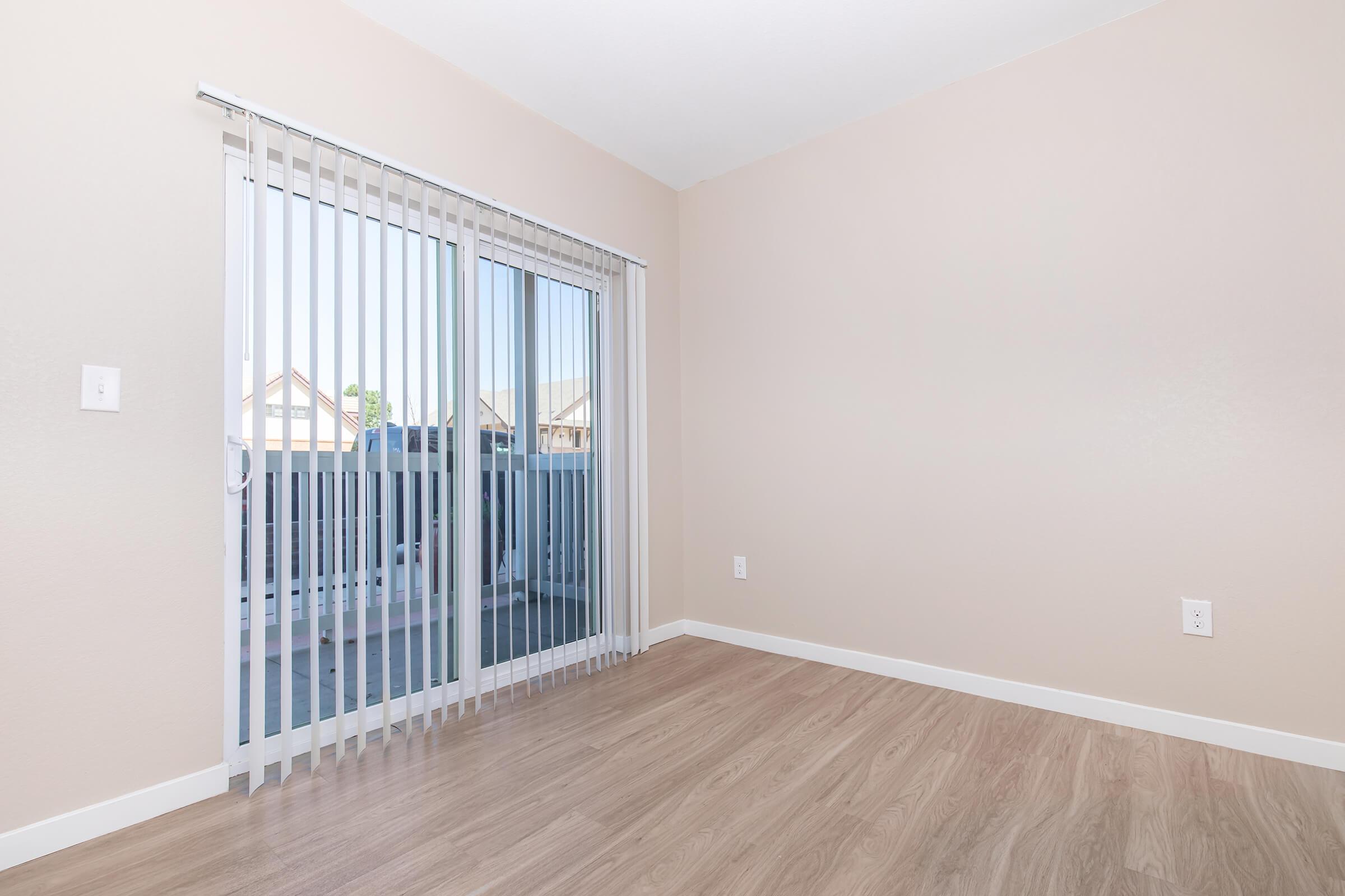 A well-lit, empty room with light beige walls and laminate flooring. There is a sliding glass door with vertical blinds leading to a small balcony outside. The space is uncluttered, lending a feeling of openness and potential for personalization.