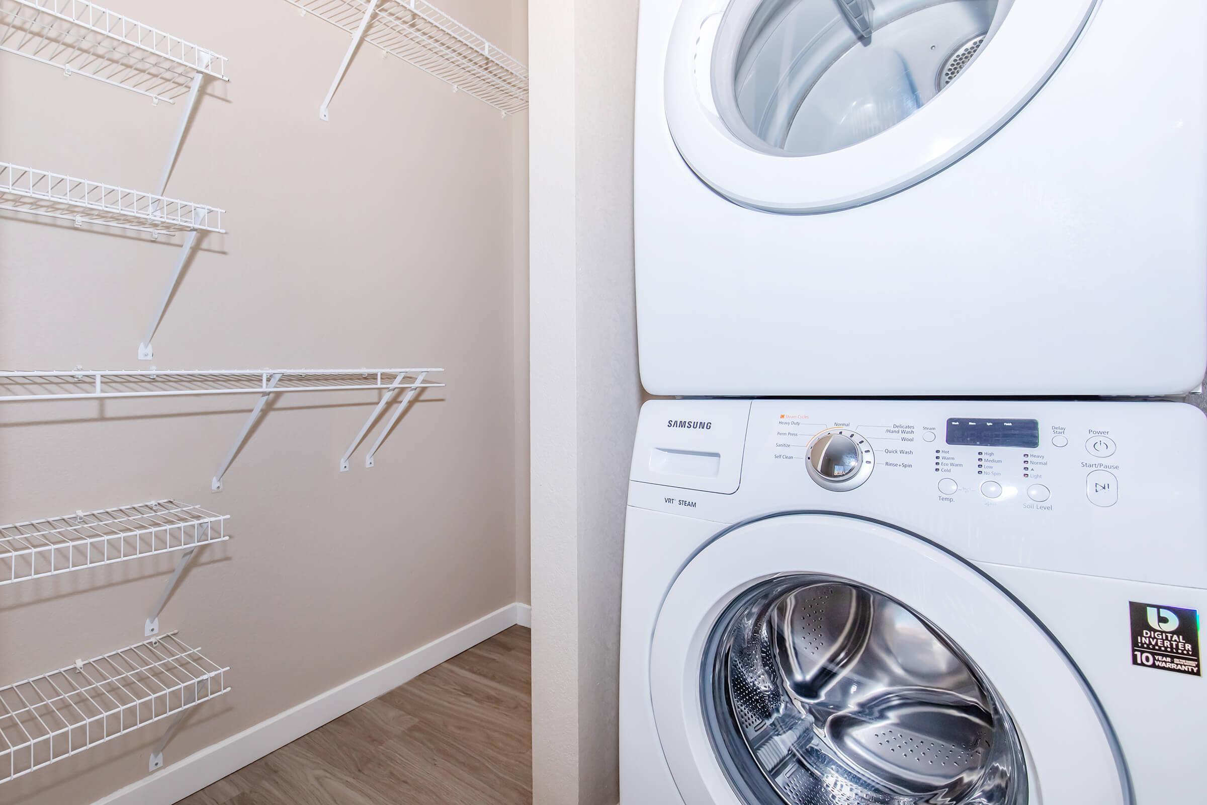 A stacked washer and dryer in a small laundry area, with several wall-mounted wire shelves next to them. The walls are a light color, and the floor is wooden laminate. The appliances are modern and white, adding to the clean and organized appearance of the space.