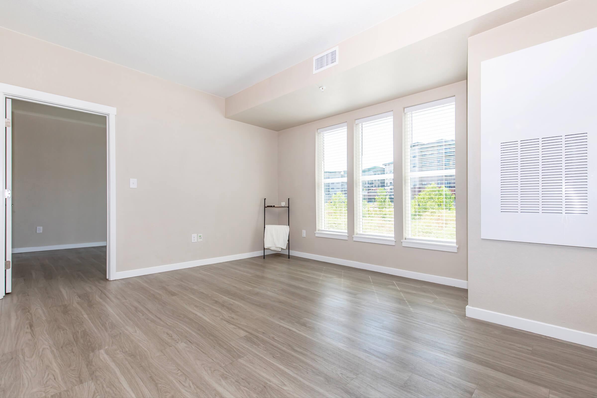 A bright and spacious room with light-colored walls and laminate flooring. It features large windows with white blinds allowing natural light to flow in. A small black rack with a towel is positioned in the corner. A doorway leads to another room, creating an open and airy atmosphere.