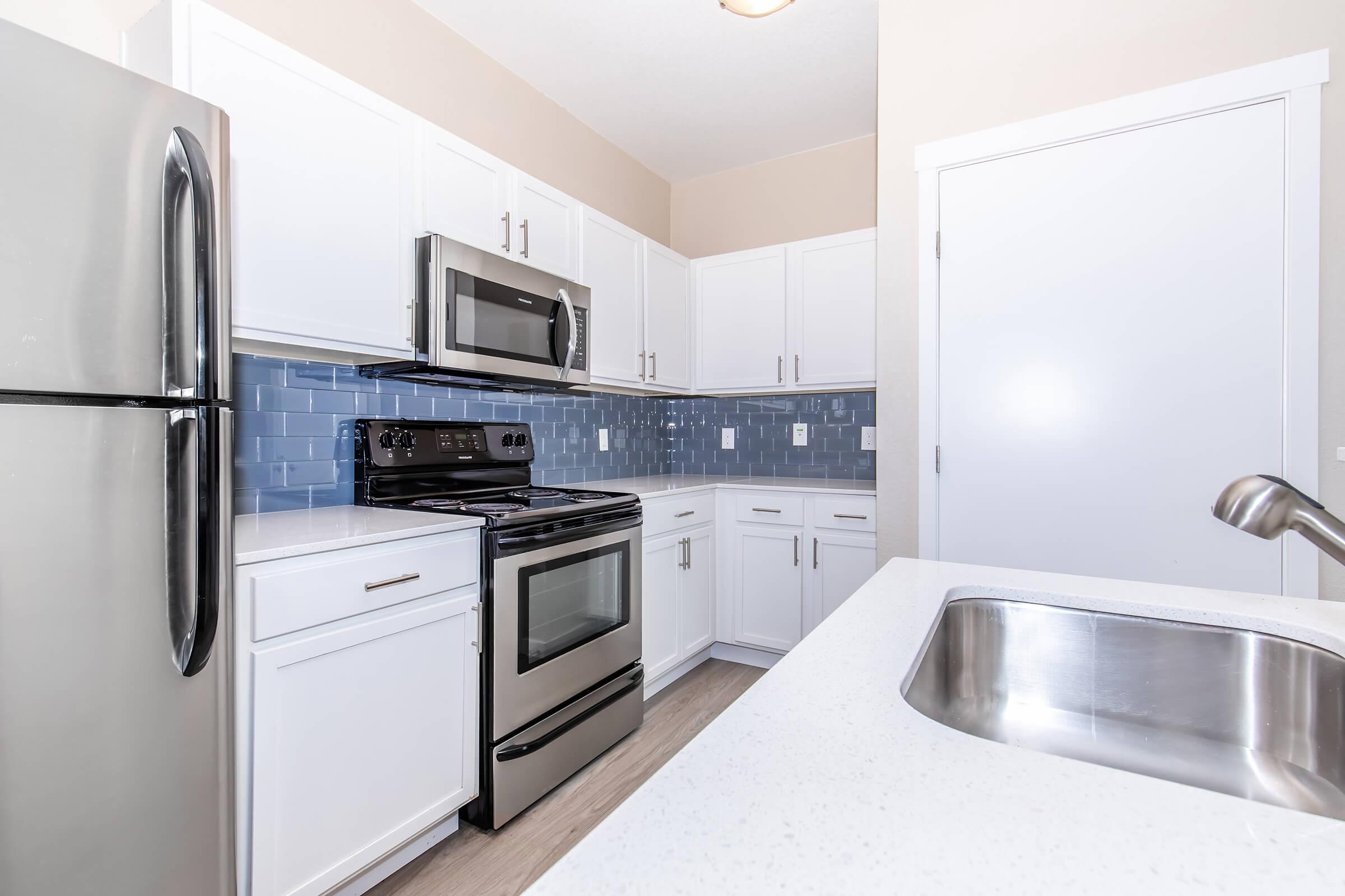 Modern kitchen featuring stainless steel appliances including a refrigerator, microwave, and oven, white cabinetry, and a light countertop. The backsplash consists of blue tiles, and the space is bright with natural light. A stainless steel sink is visible in the foreground, enhancing the contemporary design.