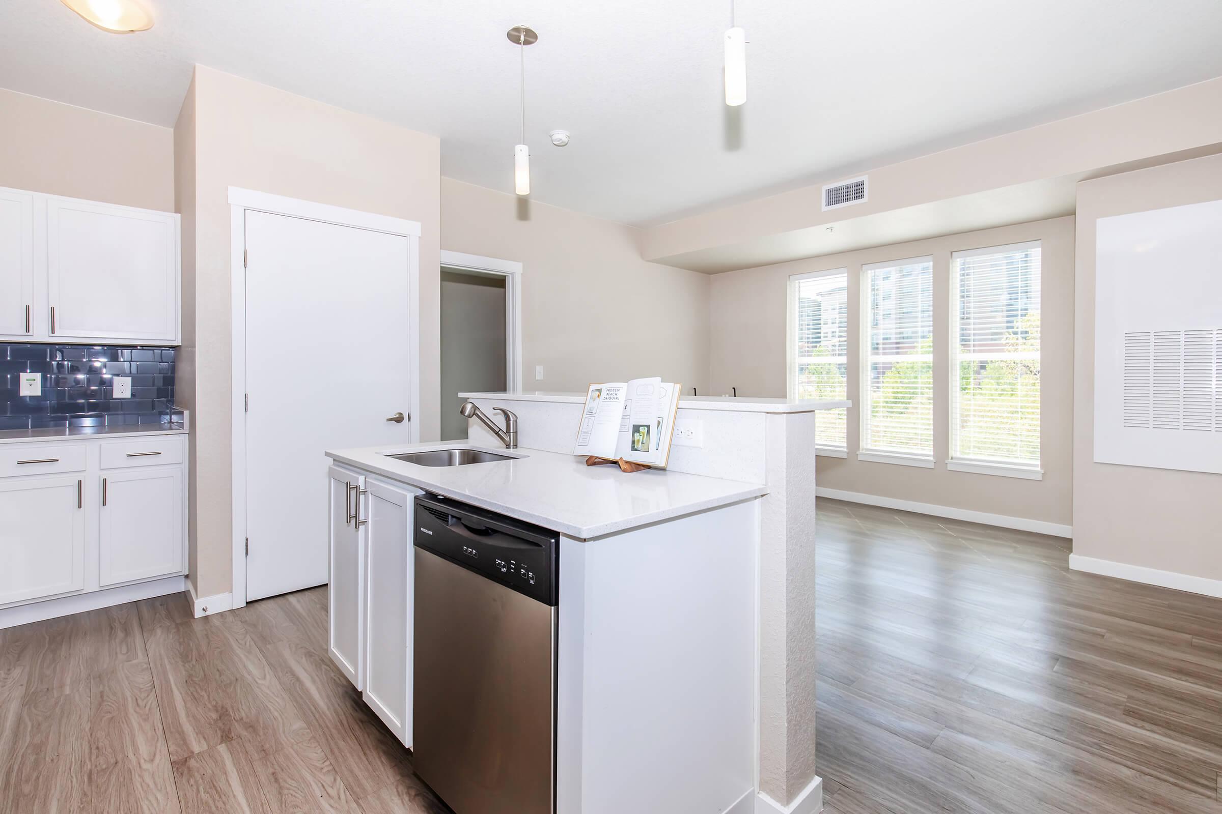 A modern kitchen featuring white cabinetry, a stainless steel dishwasher, and a dual sink. The open layout connects to a spacious living area with large windows allowing natural light. The walls are painted a soft beige, and the floor has light-colored laminate. A brochure is displayed on the countertop.