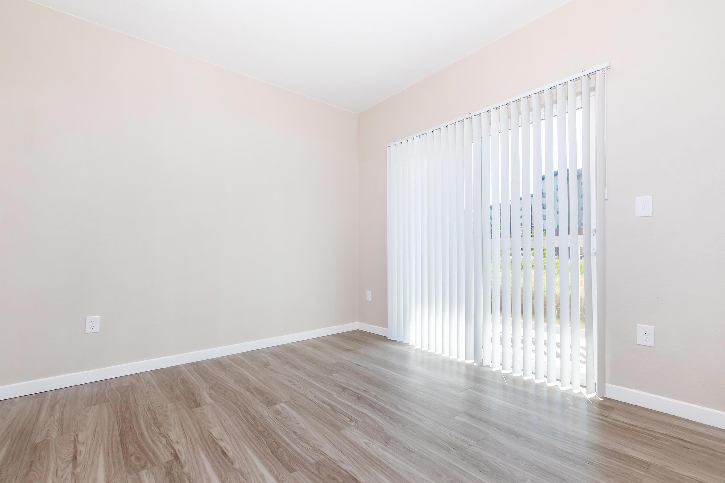 A spacious, empty room with light-colored walls and wooden flooring. There is a large window with vertical blinds allowing natural light to enter, providing a view of the outdoors. The scene conveys a clean and minimalistic aesthetic, perfect for personal customization.