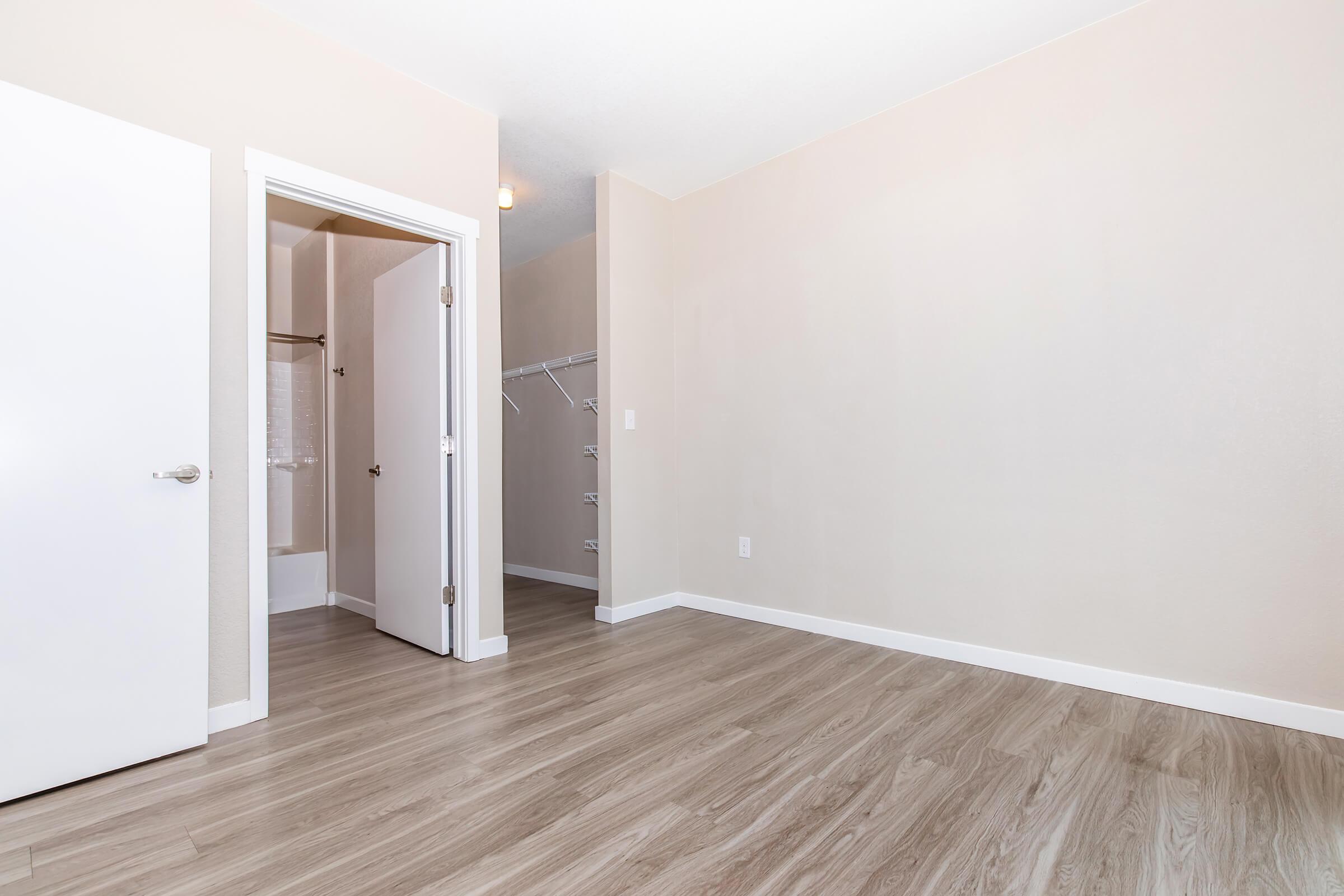 A spacious room with light-colored walls and a smooth wooden floor. There is a white door leading to a bathroom and a closet with hanging space. The room is well-lit and empty, suggesting potential for various uses, such as a bedroom or office.