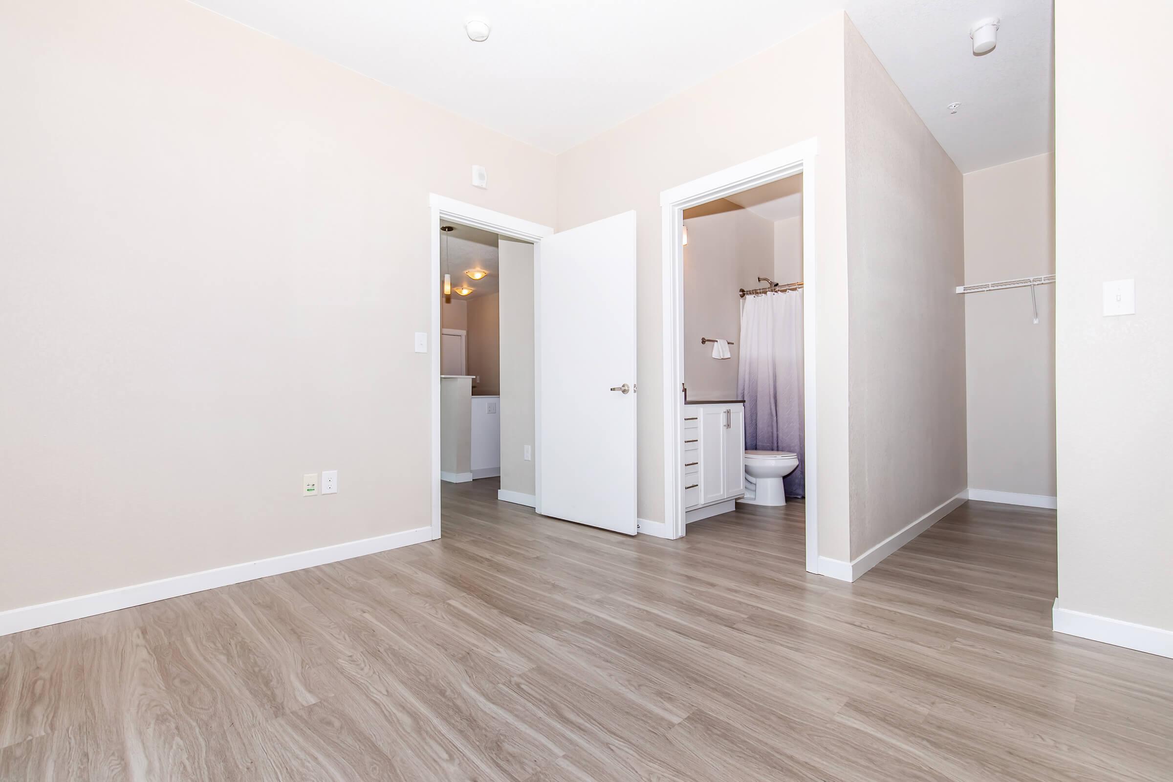 A spacious room with light-colored walls and vinyl flooring. It features two doorways: one leading to a bathroom equipped with a toilet and sink, and another opening to an adjoining area. Natural light brightens the space, creating a fresh and airy atmosphere.