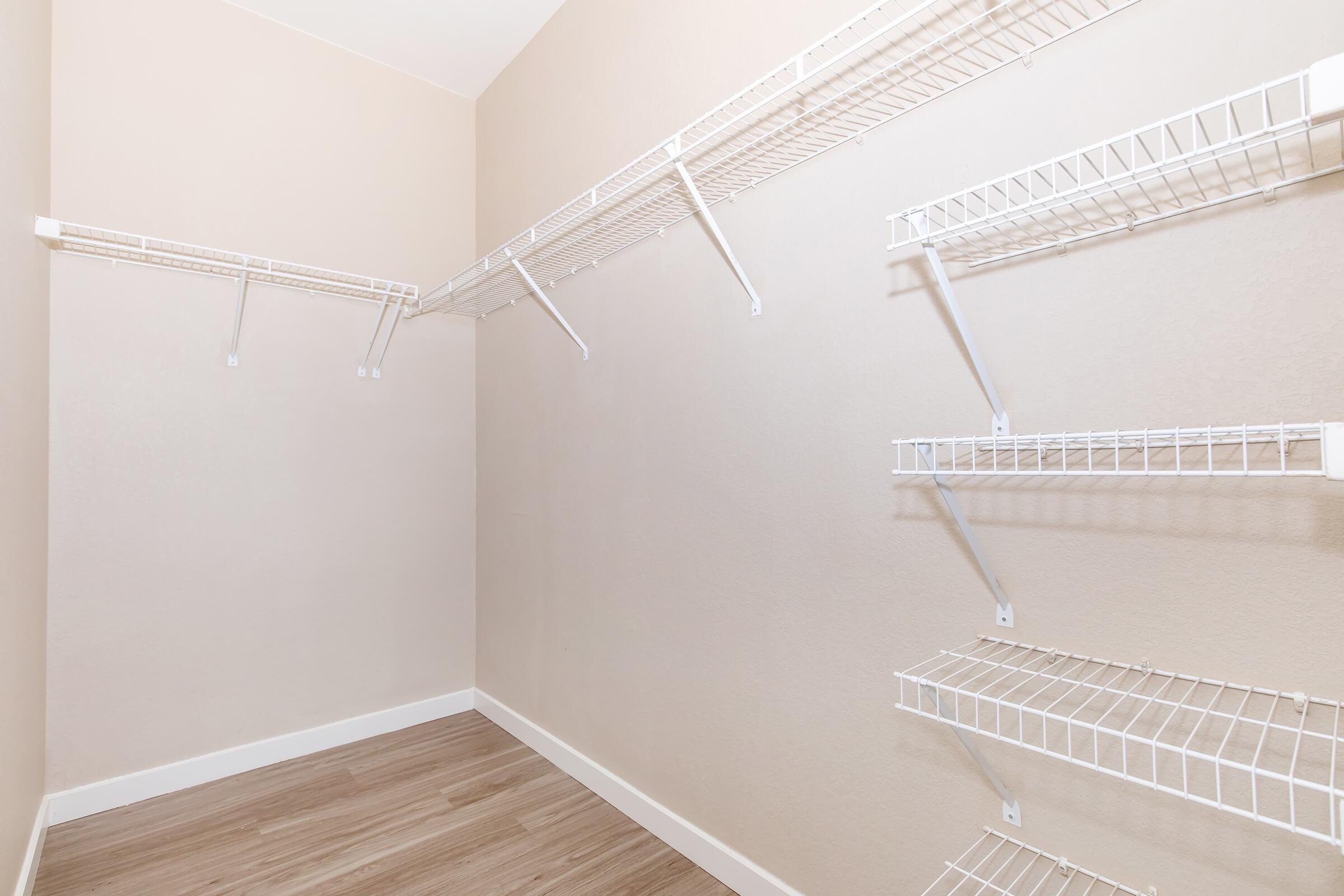 A spacious empty walk-in closet featuring white wire shelving mounted on light-colored walls, with a warm wood-like floor. The closet has an open layout, creating a bright and airy feel, ideal for organizing clothing and accessories.