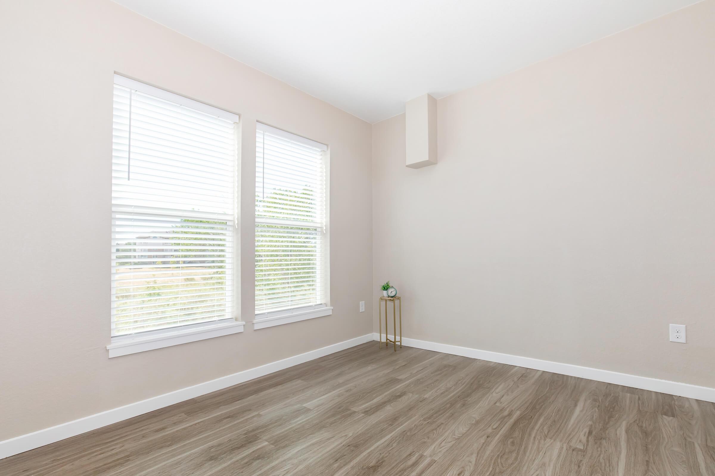 A lightly colored room with two windows featuring white blinds, allowing natural light to enter. The floor is a warm wooden laminate. In one corner, there is a small gold side table with a decorative item on top. The walls are undecorated, creating a spacious and minimalist atmosphere.