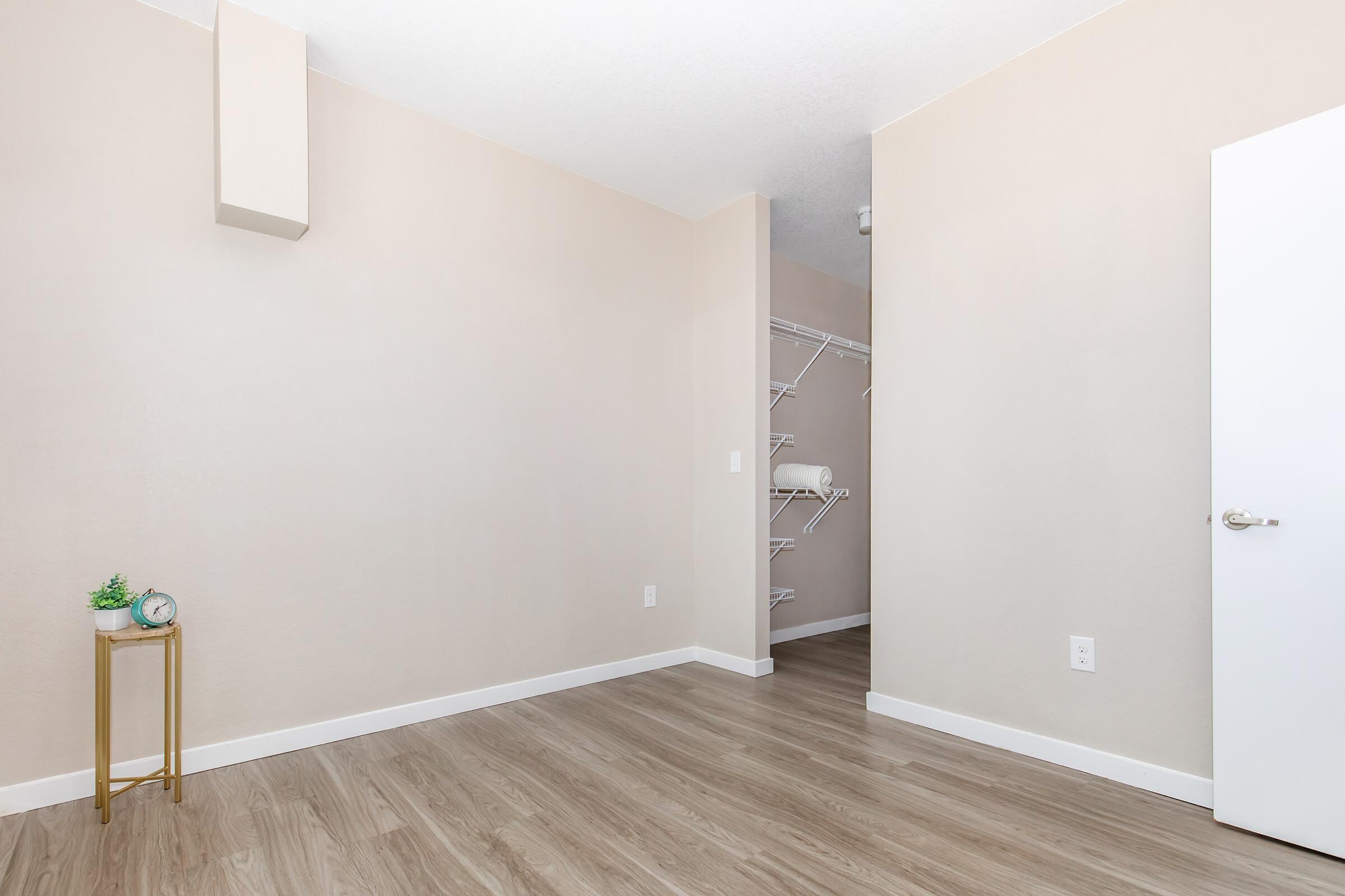 A minimalistic room with light beige walls and a wood-style floor. There is a small metallic side table with a clock and a decorative plant. A closet with open shelving is visible in the background, and a white door is on the right side of the image, indicating an exit or entry point.