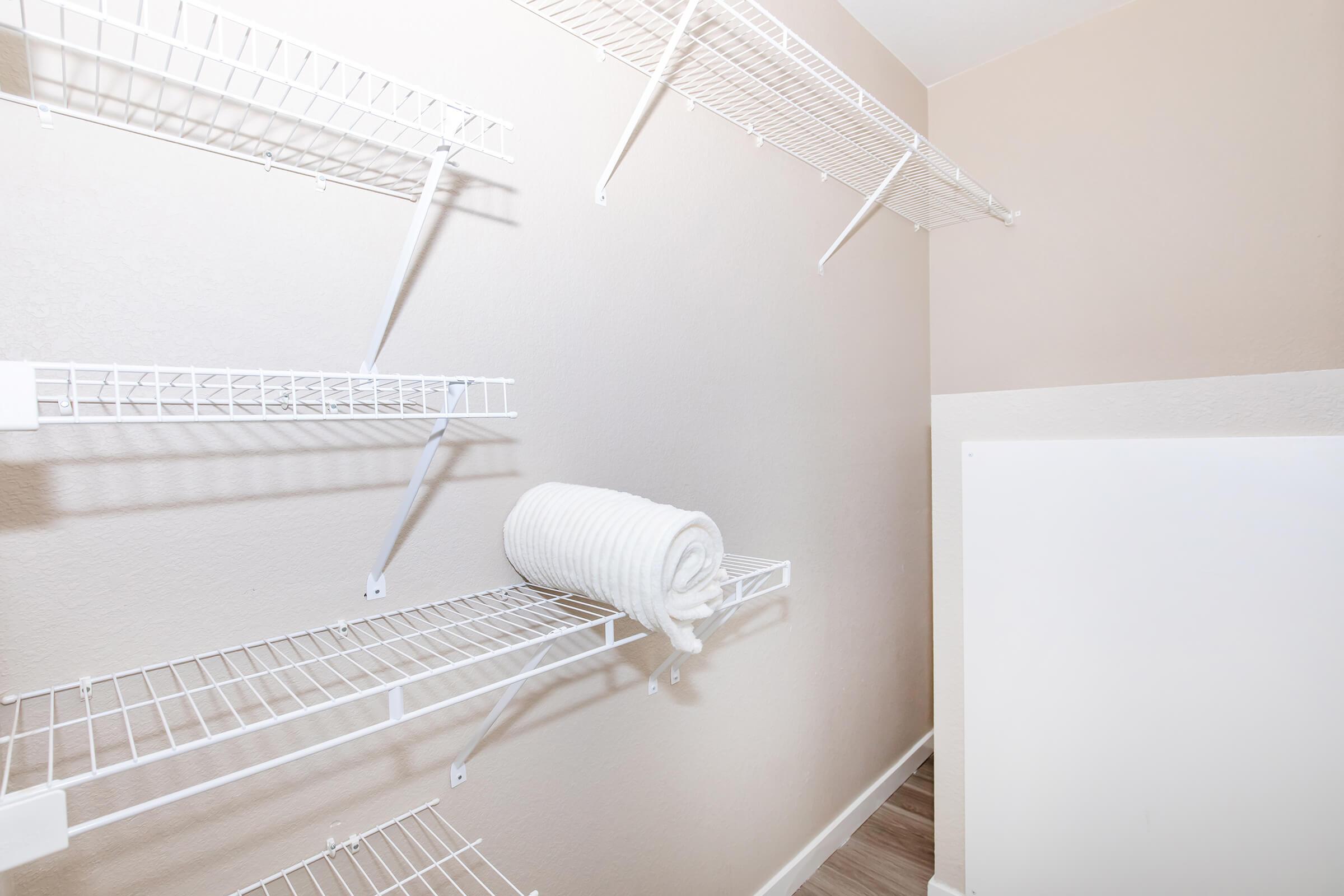 A small, empty closet with white wire shelving attached to the walls. A neatly rolled white towel is placed on one of the shelves. The walls are painted a light beige color, and the flooring appears to be a light wood or laminate finish.