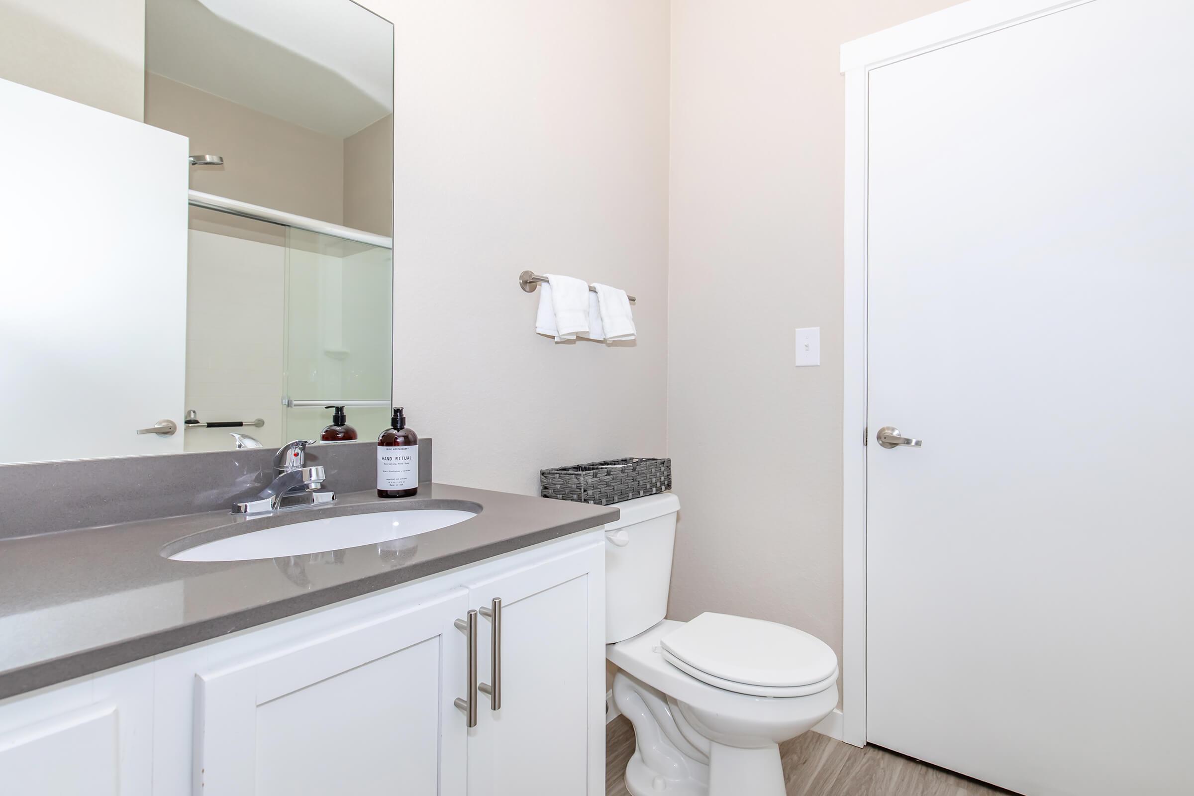 A modern bathroom featuring a white sink with a sleek faucet, a light gray countertop, a white toilet, and a door leading outside. There are towels hanging on a rack and a small basket on the counter, adding to the clean and minimalist design. Natural light brightens the space through a large mirror.