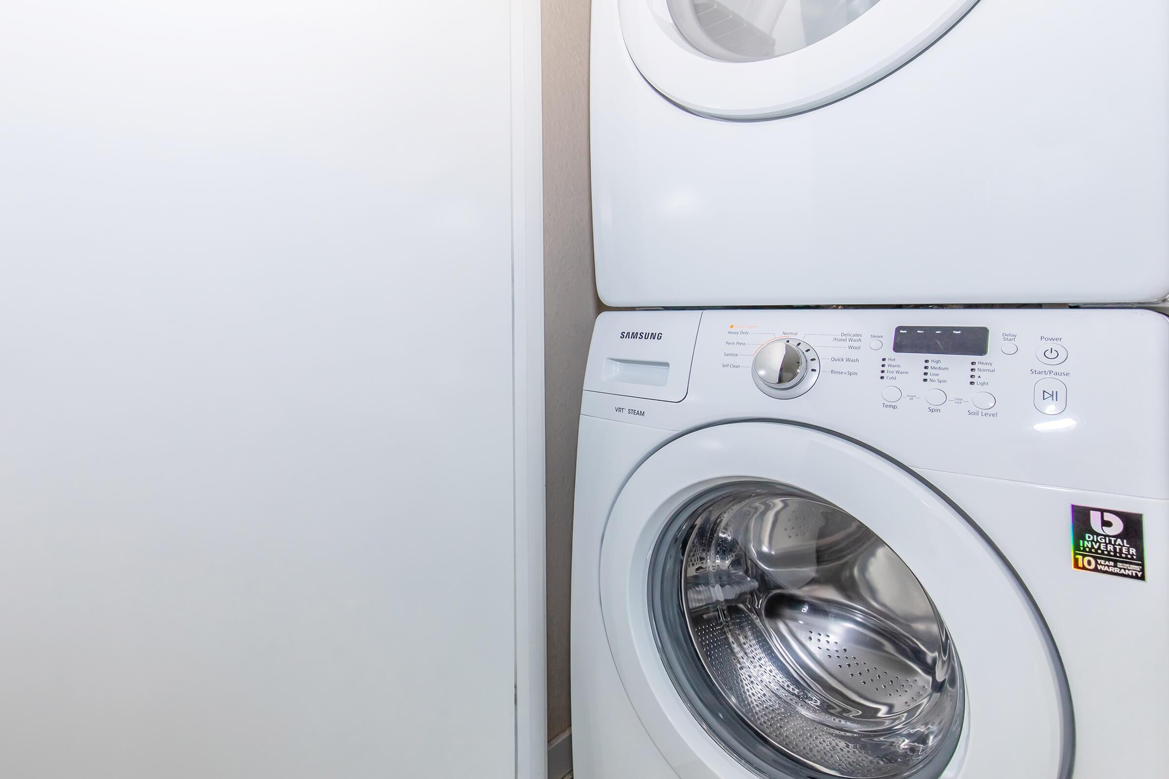 A stacked white Samsung washing machine and dryer in a laundry space. The washing machine features a control panel with various settings, and the dryer is positioned directly above it. A light gray wall serves as the background, with a portion of a white cabinet visible to the left.