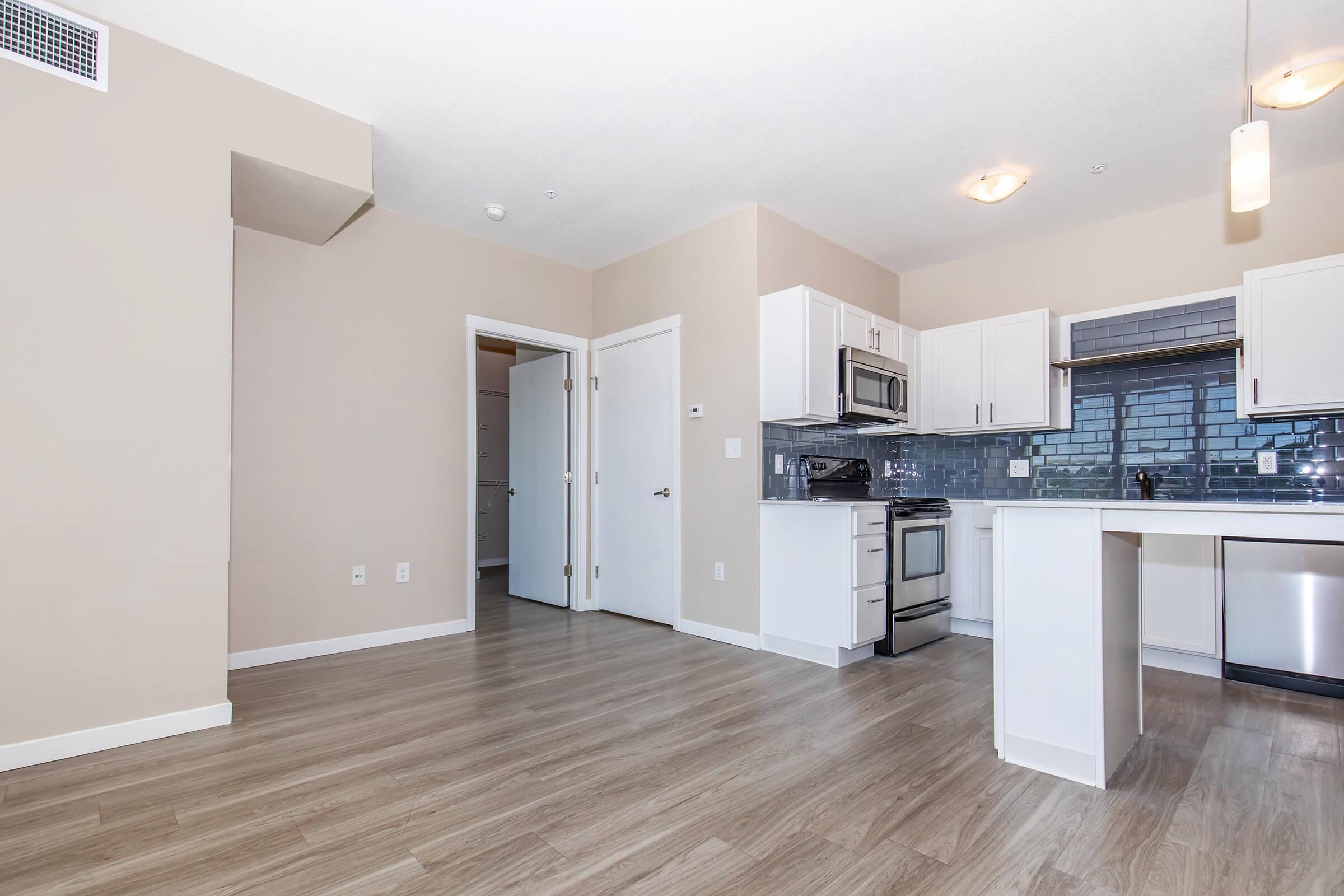 A modern kitchen and living space featuring light wood flooring, white cabinetry, stainless steel appliances, and a blue tile backsplash. An open layout leads to a doorway and hallway on the left, with ample natural light from surrounding windows. The walls are painted in a neutral color.