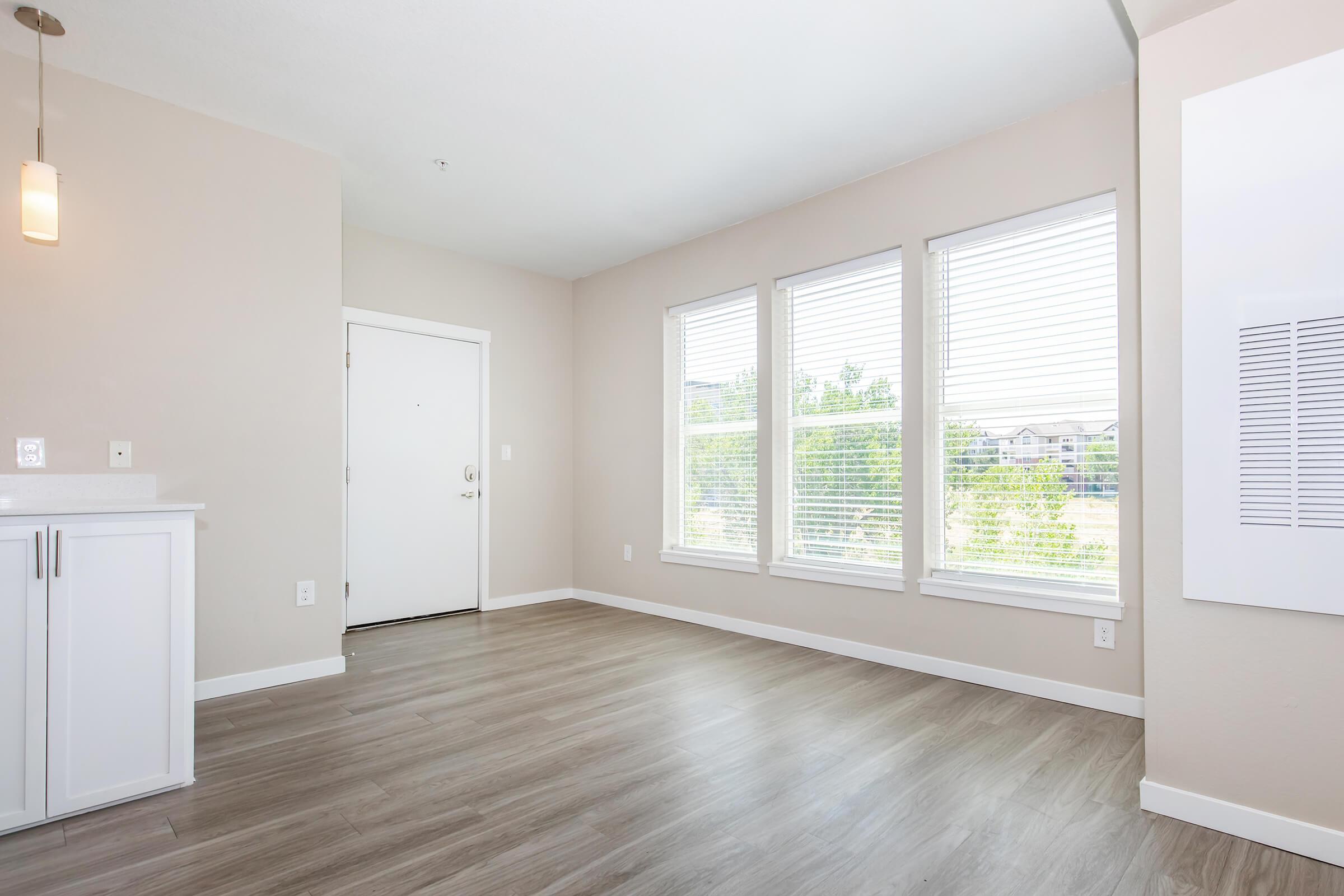 A bright, vacant room featuring light-colored walls and large windows with blinds. The floor is covered with light wood laminate. There is a white entry door on the left and a white cabinet with a countertop on the left side. The room has a modern, open feel, with natural light illuminating the space.