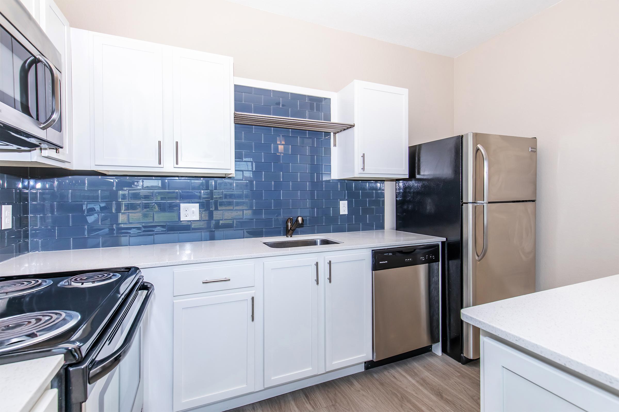 Modern kitchen featuring white cabinets, stainless steel appliances including a refrigerator and dishwasher, a black stove with an oven, and a blue tile backsplash. The countertops are light-colored, and the flooring is a neutral wood laminate. Abundant natural light enhances the spacious feel.