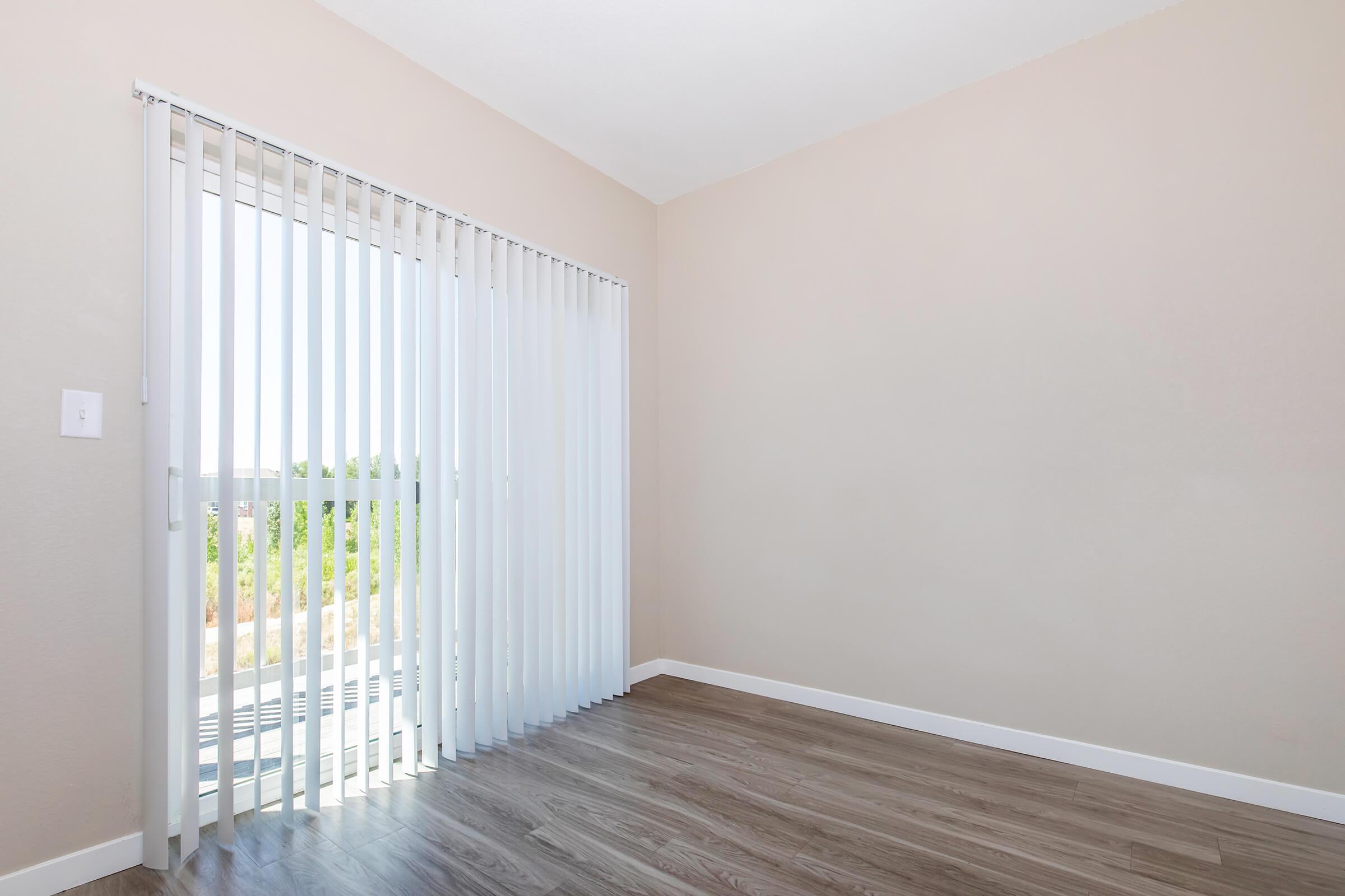 A bright, empty room with light beige walls and a large sliding glass door featuring white vertical blinds. The floor is covered in a warm wood-like laminate, and through the blinds, green foliage is visible outside, suggesting a serene view. The space has a clean and modern aesthetic.