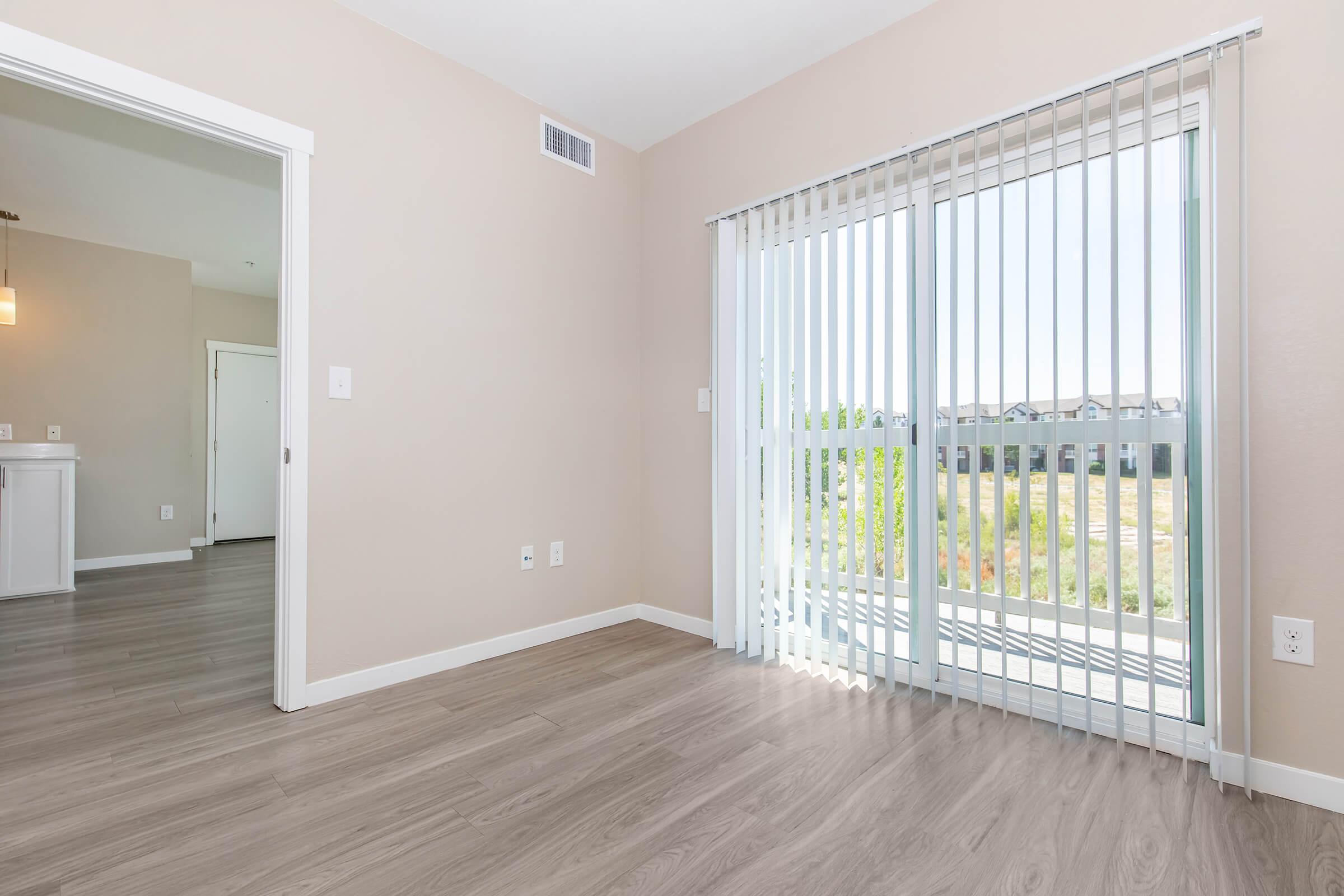 A spacious, well-lit room featuring light-colored walls and a wood-textured floor. Large sliding glass doors with vertical blinds open to a view of greenery outside. Natural light fills the space, enhancing the open, inviting atmosphere. An adjoining room is partially visible in the background.