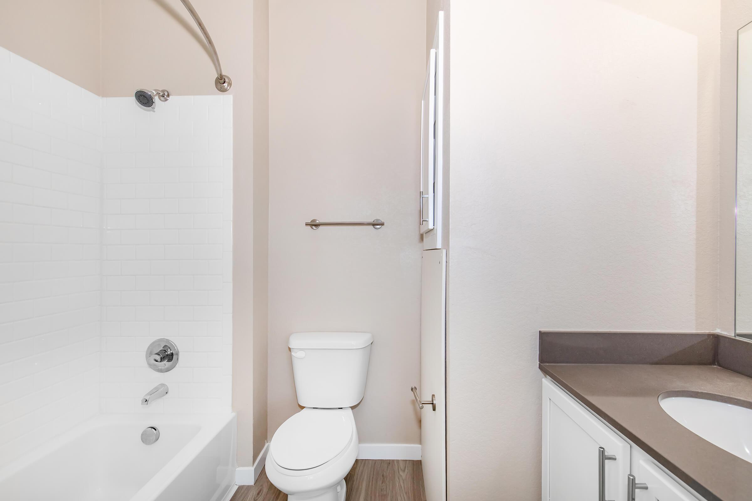 A clean and modern bathroom featuring a white bathtub with a shower, a toilet, and a vanity with a sink. The walls are painted in a soft beige color, and there is a window allowing natural light to enter. The flooring is a light, wood-like material.