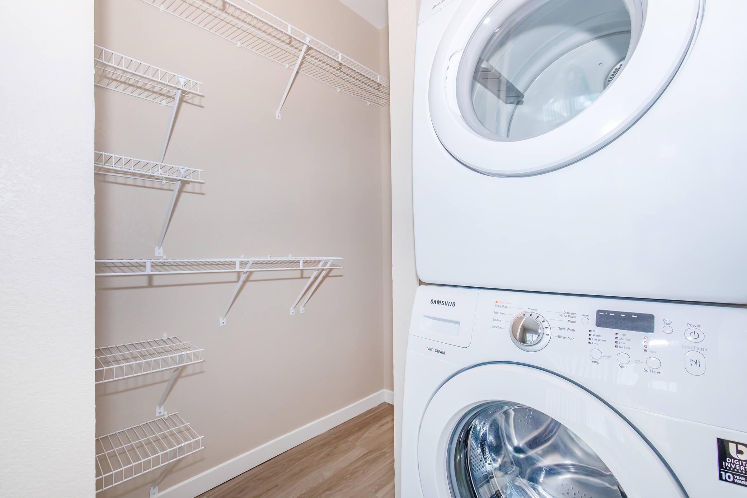 A laundry area featuring a stacked washer and dryer in white, positioned next to a beige wall. Wire shelving units are mounted above the appliances, providing storage space. The floor is finished with light wood laminate, creating a clean and modern look.