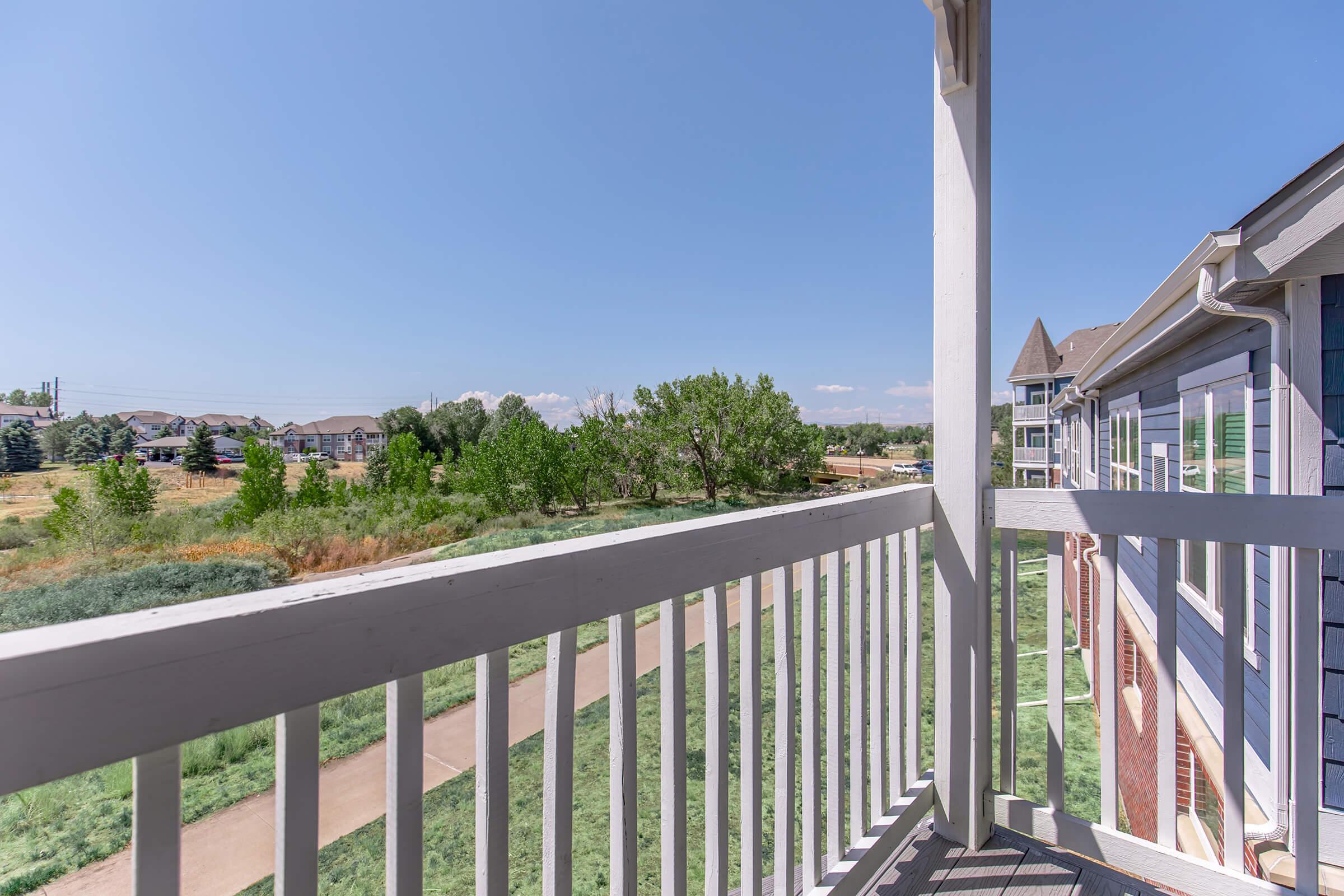 A view from a balcony showcasing a blue sky with a few clouds, green trees, and a pathway lined with grass. In the distance, residential buildings can be seen, creating a peaceful suburban landscape. The railing of the balcony is in the foreground, framing the scene.