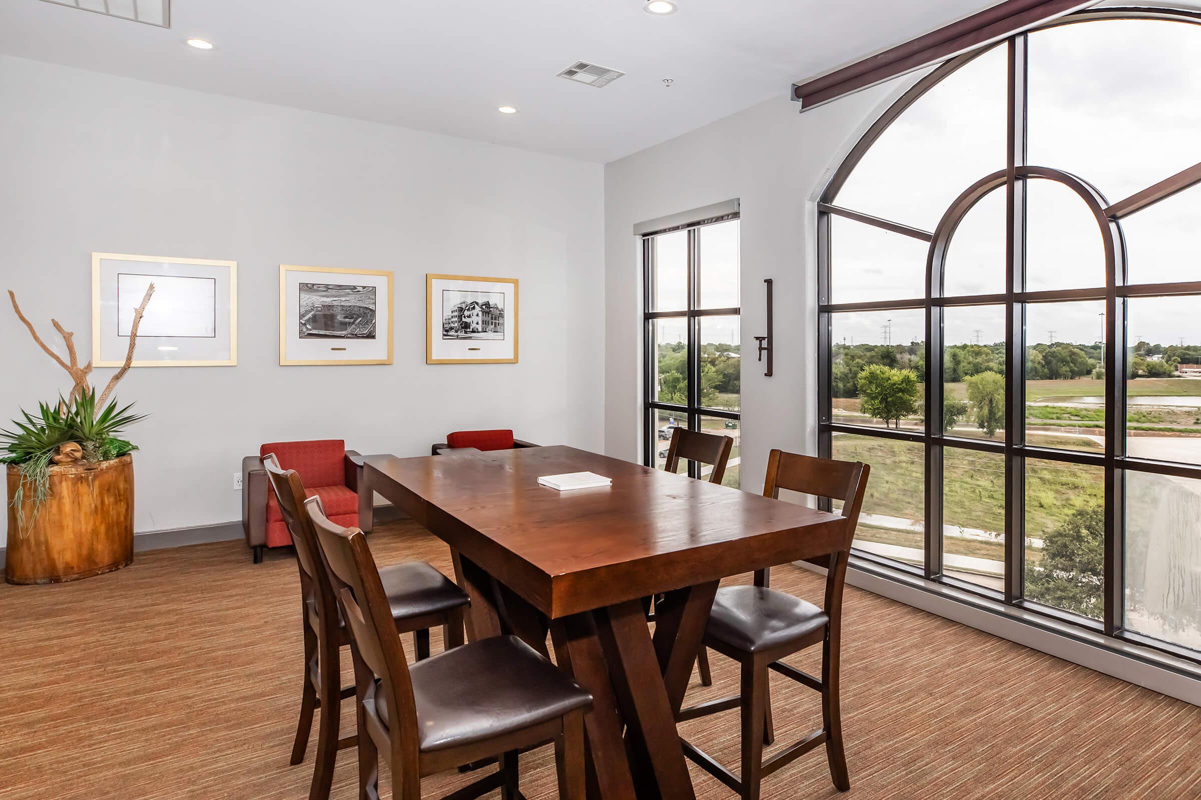 A modern meeting room featuring a large wooden table surrounded by four chairs. There are three framed photographs on the wall, and a decorative plant in a wooden planter. The room has a large arched window offering a view of greenery outside, with natural light flooding the space.