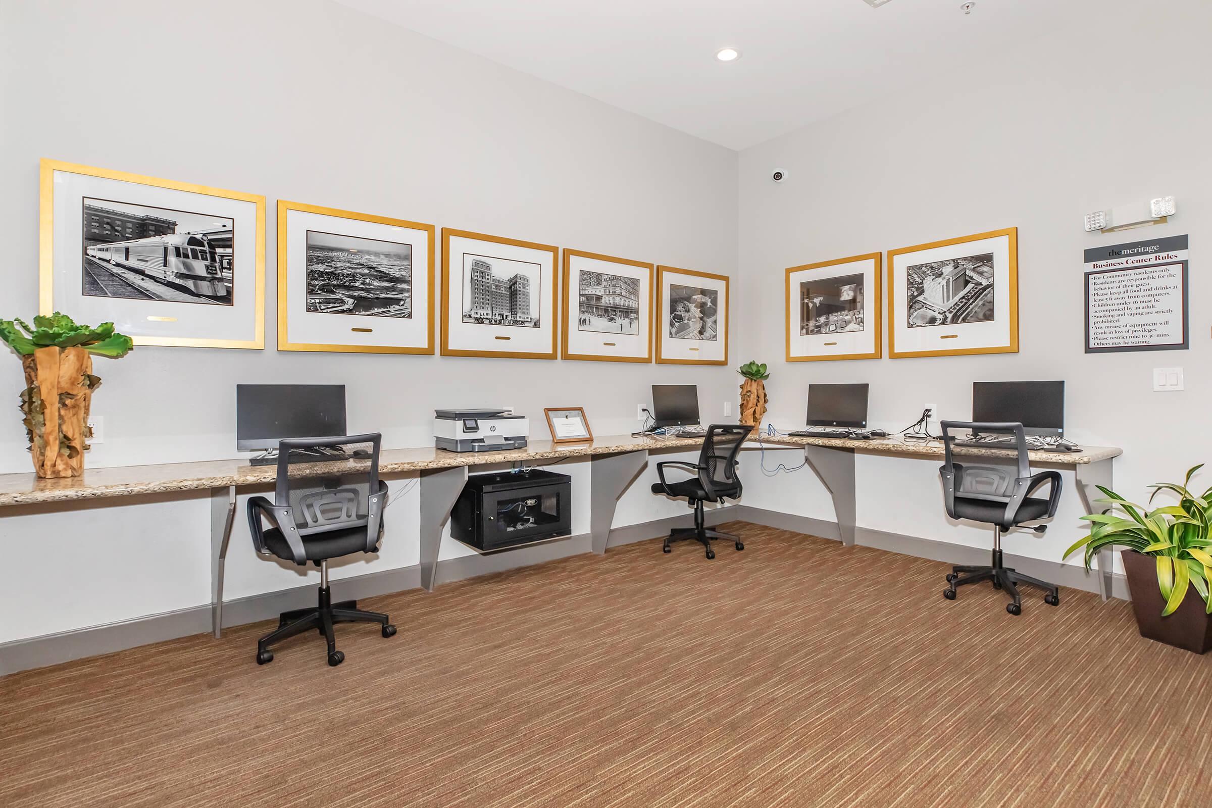 A modern workspace featuring a beige carpet, four black office chairs, and computers at three desks. The walls are adorned with framed black-and-white photographs in gold frames, alongside a bulletin board and a printer. Potted plants add a touch of greenery to the environment.