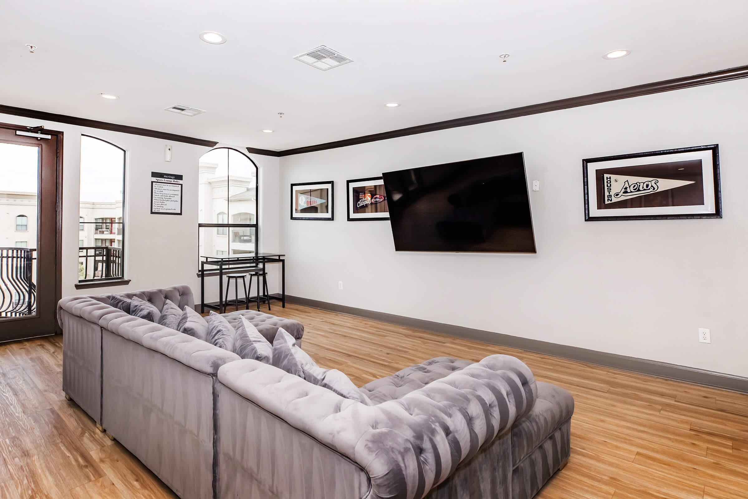 A modern living room featuring a plush gray sectional sofa, a mounted flat-screen TV on the wall, framed artwork, and a small dining table by the window. The room has wooden flooring and large windows, allowing for natural light to fill the space.