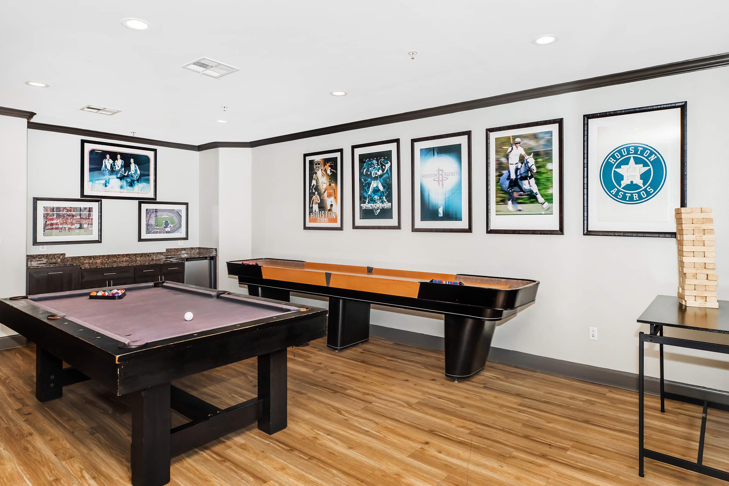 A game room featuring a billiard table, an air hockey table, and a Jenga game stacked beside it. The walls are adorned with framed sports posters, including football and baseball themes. The room has wooden flooring and modern lighting, creating a lively atmosphere for entertainment.