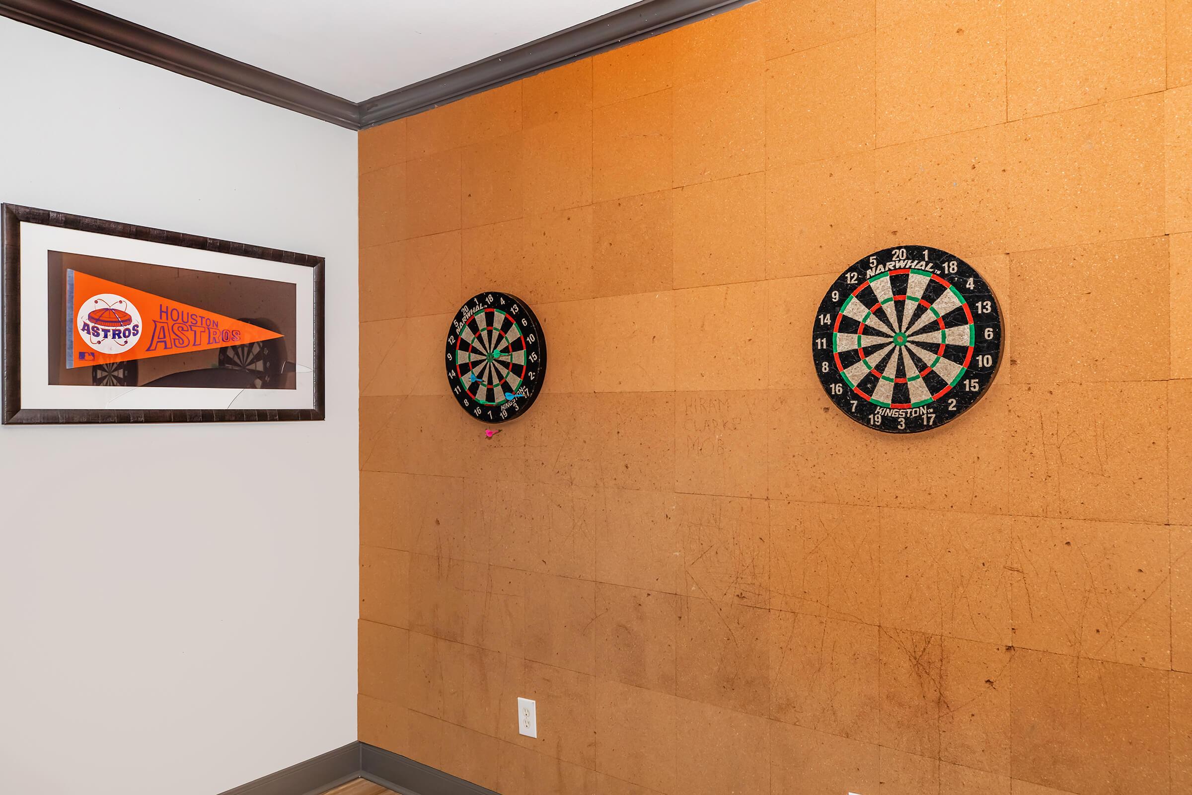 Two dartboards mounted on a cork wall, one slightly higher than the other. A framed Houston Astros pennant is displayed to the left, adding a sports-themed decor element to the room. The overall color scheme is warm with earthy tones.