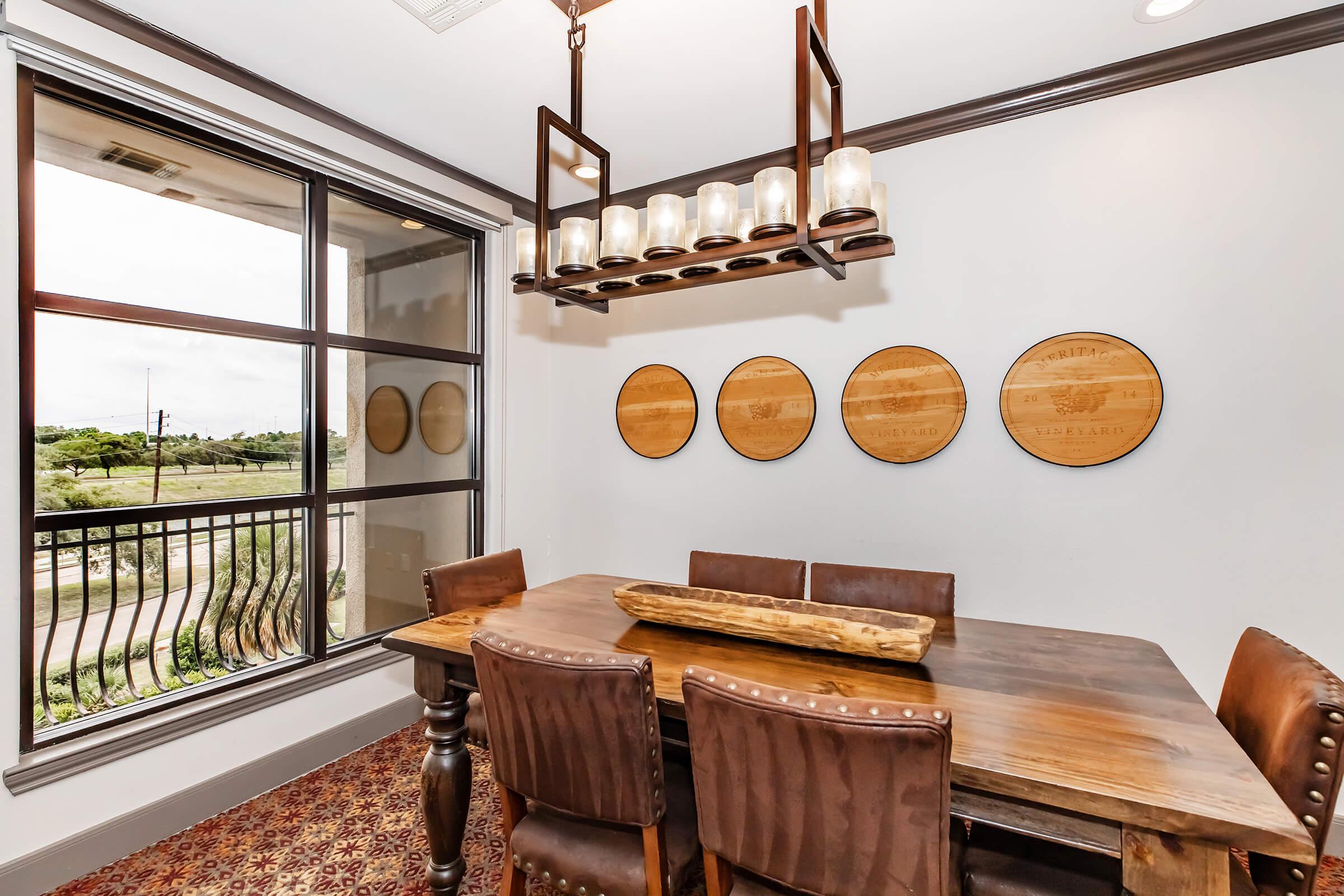 A dining area featuring a wooden table surrounded by upholstered chairs. A modern chandelier with candles hangs above the table. On the wall, there are four round wooden decorations. A large window offers a view of the outdoors, allowing natural light to fill the space.