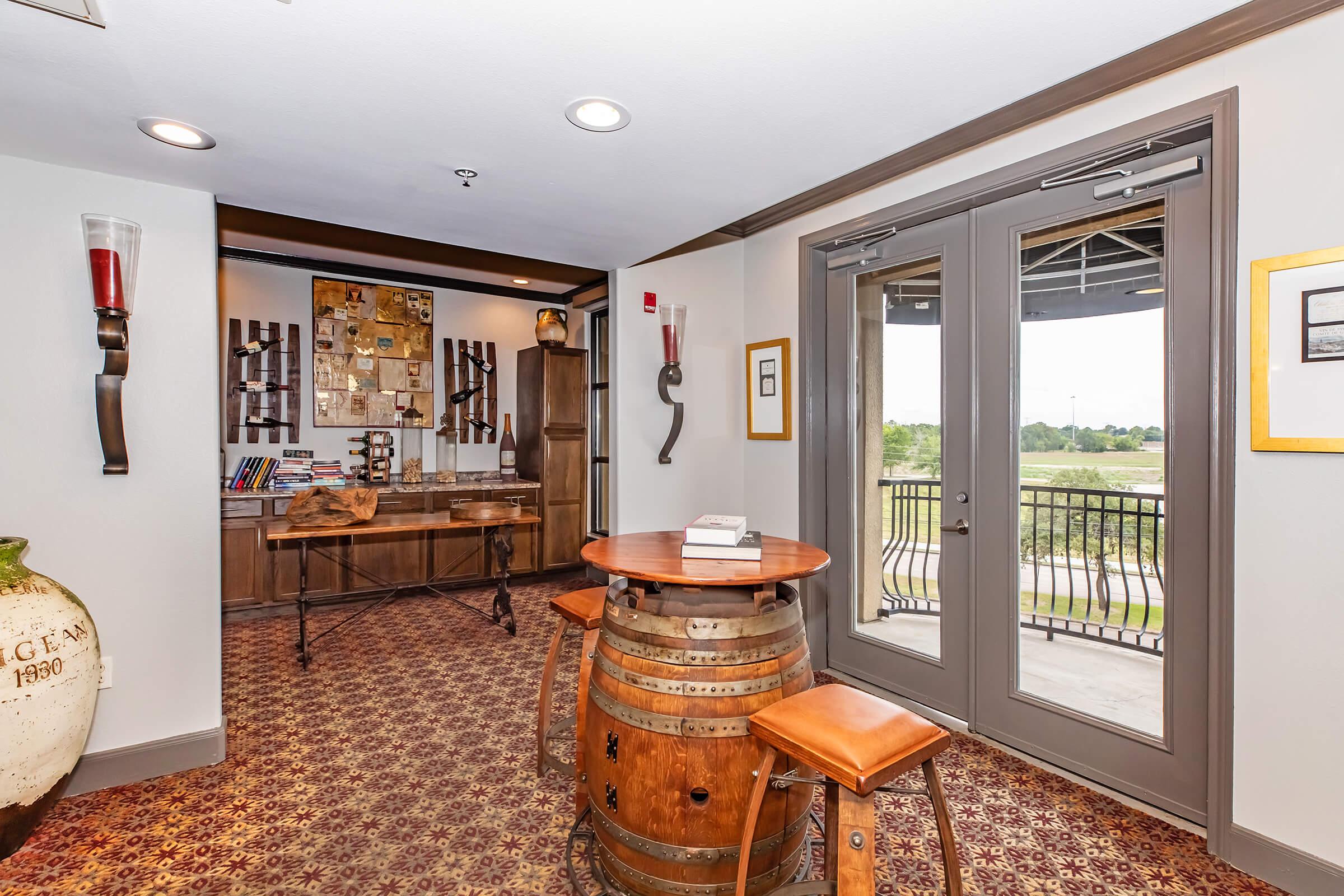 A cozy, well-decorated room featuring a wooden barrel table and two stools. It has large glass doors leading to a balcony, adorned with artwork and decorative items, against a backdrop of warm colors and textured walls. Natural light fills the space, creating an inviting atmosphere.