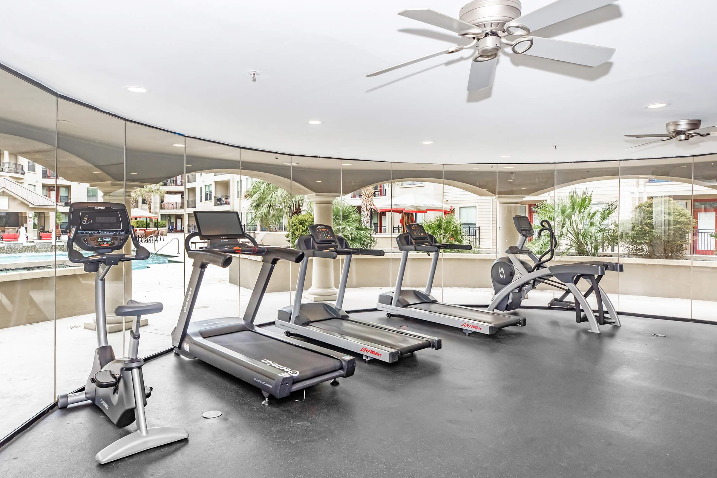 A modern gym area featuring several exercise machines including treadmills and an elliptical, situated in a brightly lit space with large windows. The gym overlooks an outdoor area with palm trees and a pool. The ceiling has a fan for ventilation, creating a comfortable workout environment.