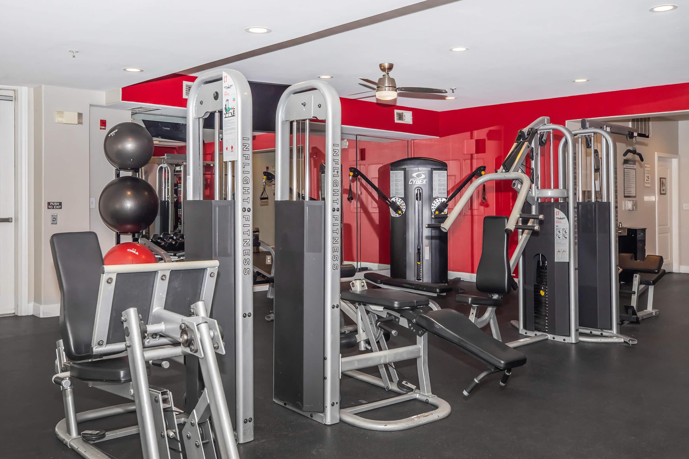A modern gym interior featuring various workout equipment, including weight machines and benches. The space is brightly lit with red and white walls, and has a clean, organized layout. Exercise balls are placed in the corner, contributing to a motivational workout environment.