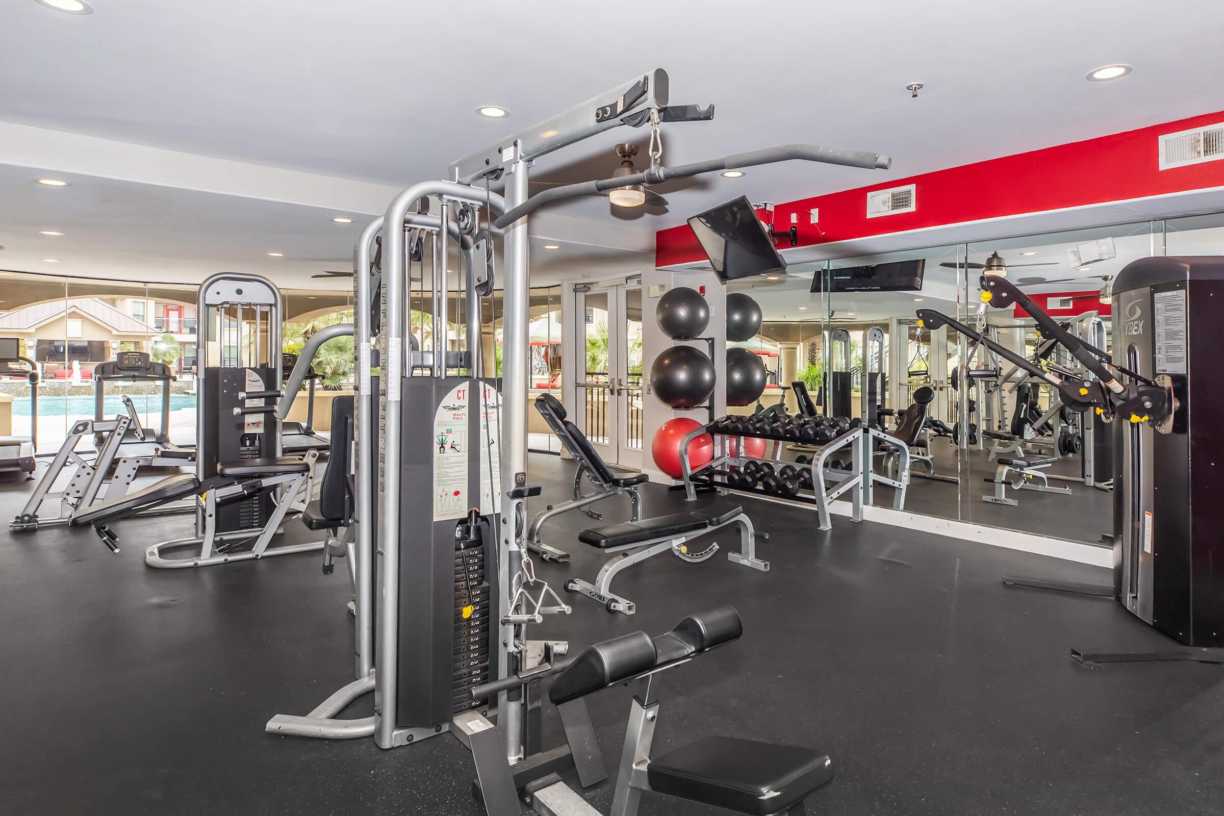 A well-equipped gym featuring various workout machines, free weights, and exercise balls. The space has a mirrored wall and bright lighting, creating an inviting atmosphere for fitness activities.