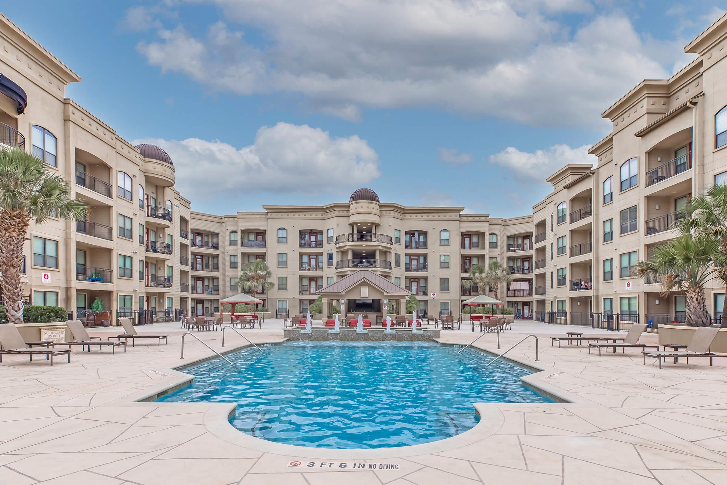 A spacious apartment complex featuring a central swimming pool surrounded by lounge chairs, palm trees, and shaded seating areas. The building has multiple stories with balconies and decorative architecture under a cloudy sky. Clear blue water reflects the surroundings.
