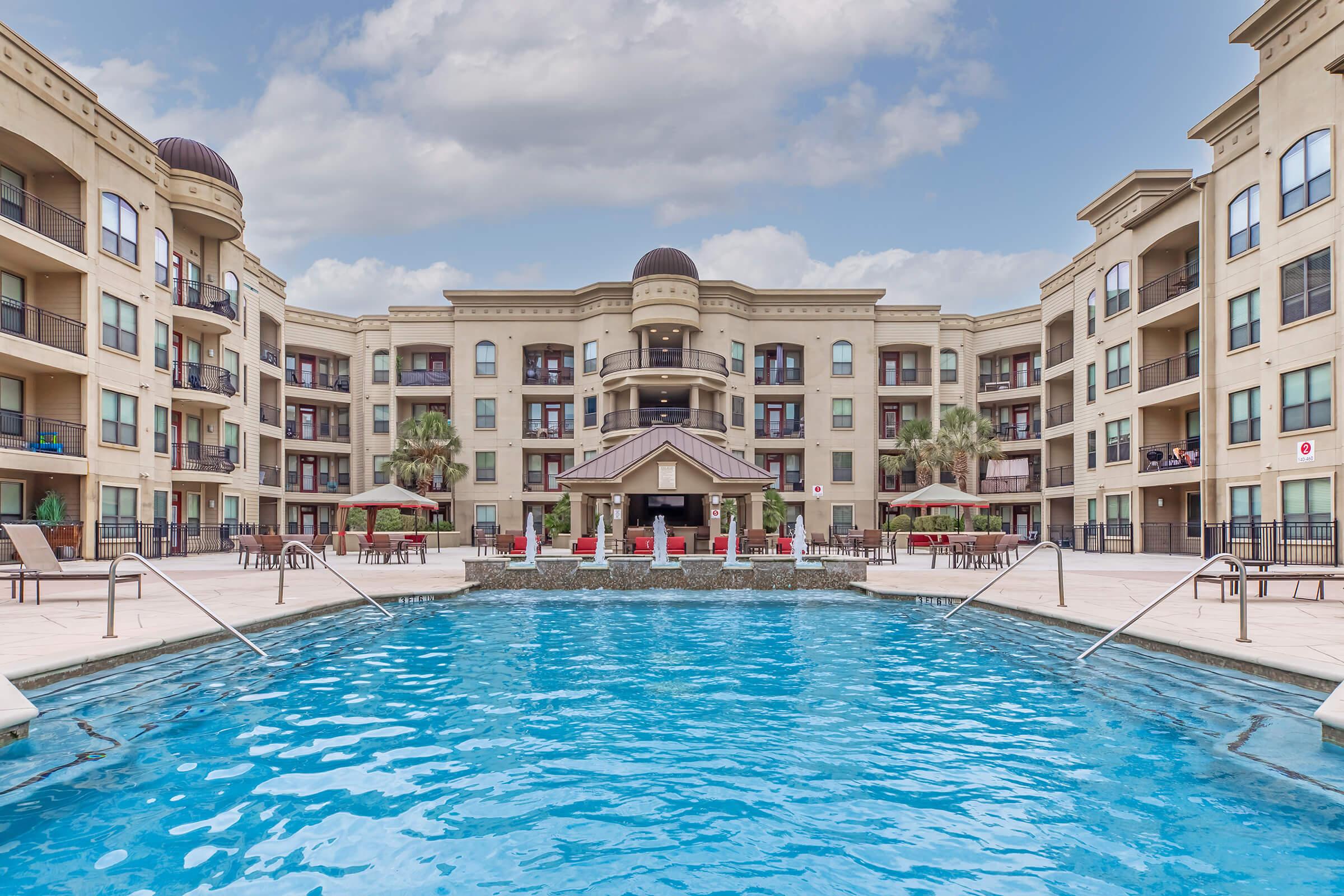 A luxury apartment complex featuring a large swimming pool in the foreground. The building has multiple stories, with balconies visible on either side. The pool area has lounge chairs and tables, surrounded by palm trees, under a partly cloudy sky.
