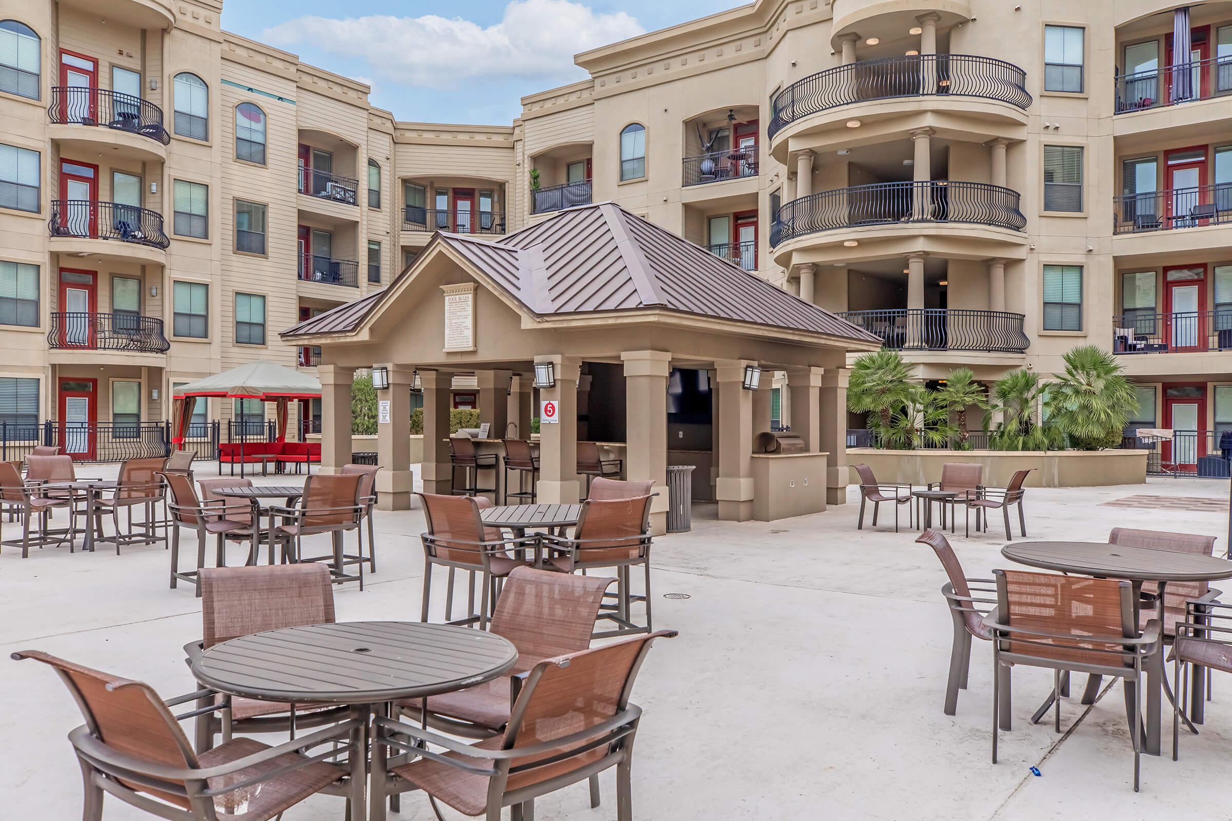 A landscaped courtyard featuring a pavilion and several outdoor tables and chairs. Surrounding the area are multi-story apartment buildings with balconies. Lush greenery adds to the inviting atmosphere, while a shaded seating area provides a space for relaxation. The sky is partly cloudy.