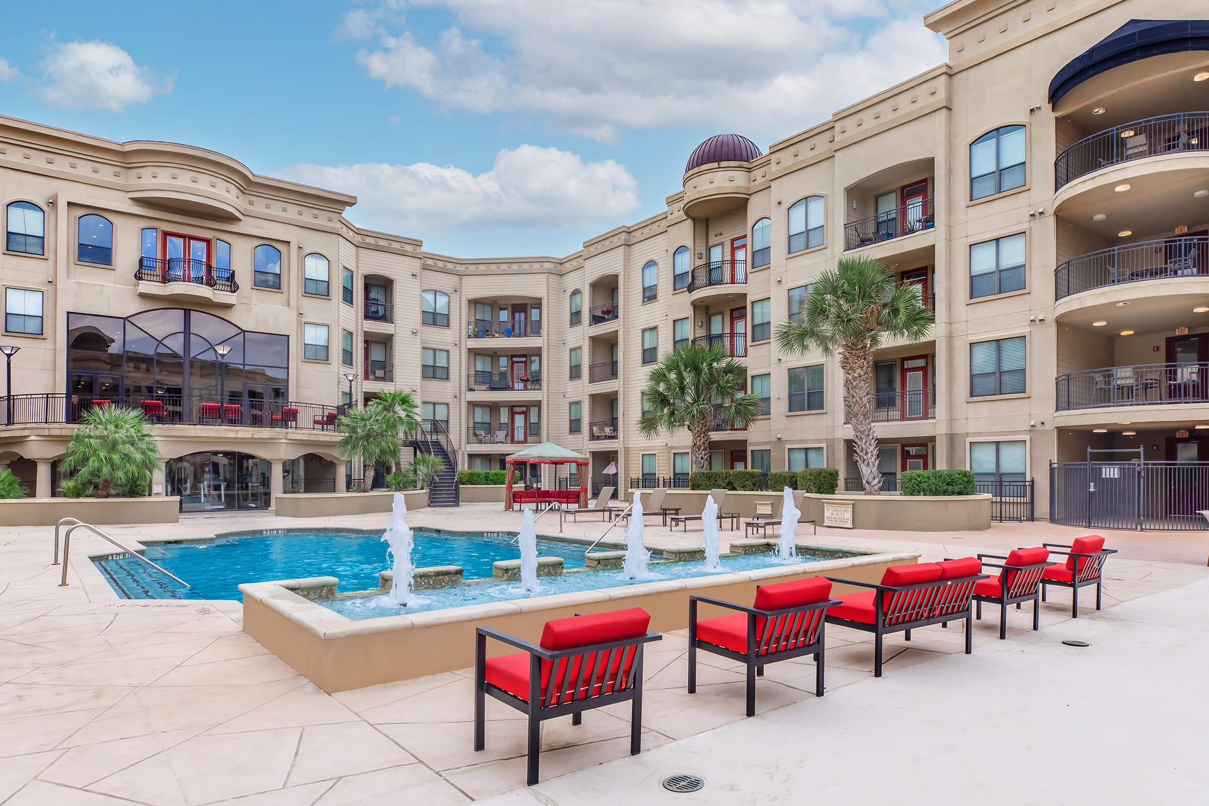 A spacious apartment complex courtyard featuring a blue swimming pool with fountains, surrounded by palm trees. Red lounge chairs are arranged around the pool area, and the building showcases modern architecture with balconies and large windows under a blue sky with scattered clouds.
