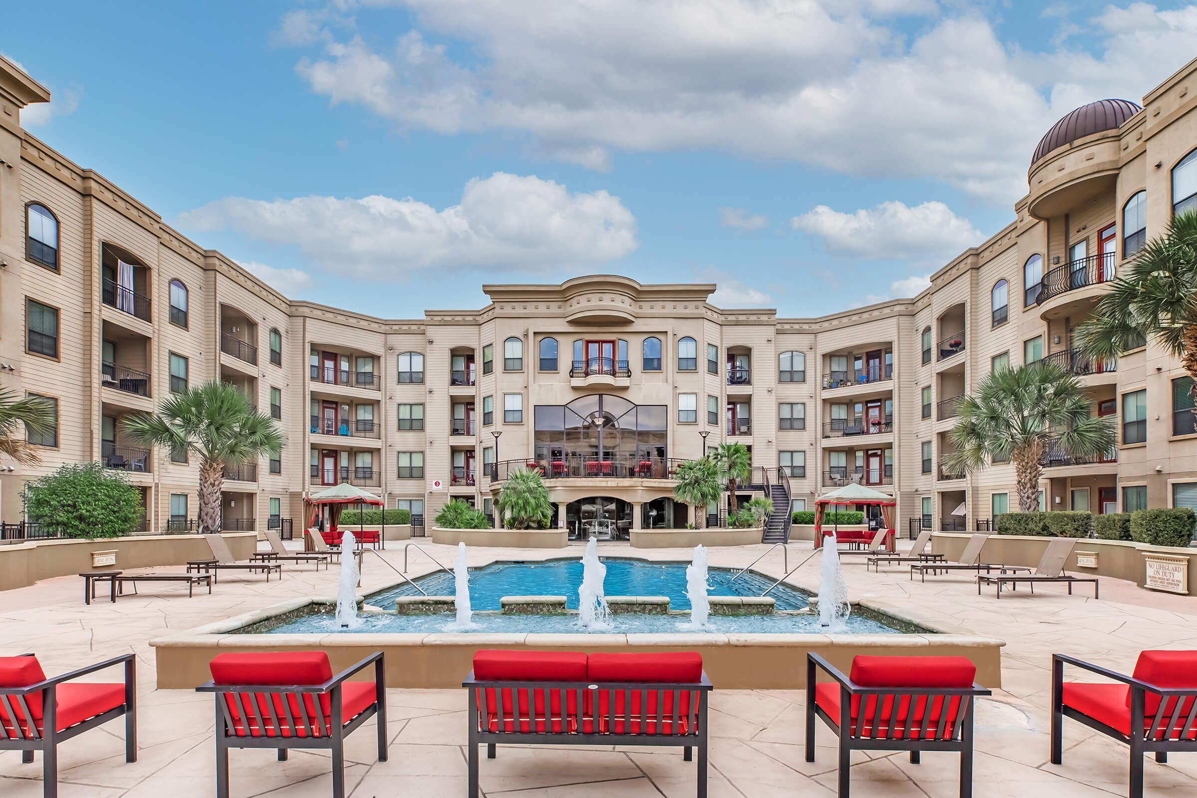 A serene apartment complex courtyard featuring a swimming pool with fountain jets. Surrounding the pool are red lounge chairs and landscaped greenery, with elegant buildings showcasing balconies in the background against a cloudy sky.