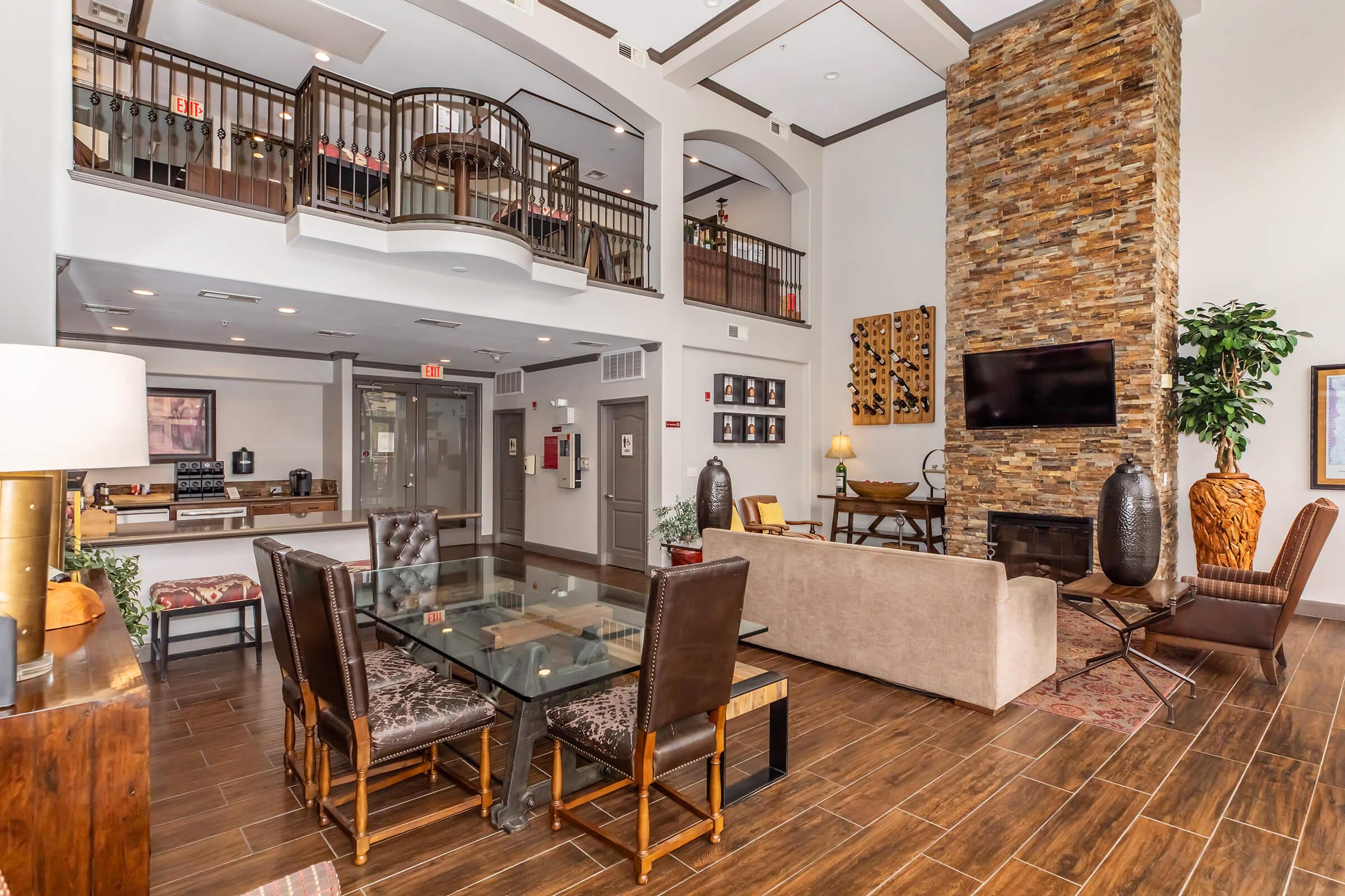 A stylish modern lobby featuring a seating area with a beige sofa, dark leather chairs, and a glass dining table. A stone accent wall adds texture, and a large flat-screen TV is mounted above a fireplace. The space includes decorative art pieces, plants, and a well-lit reception area in the background.