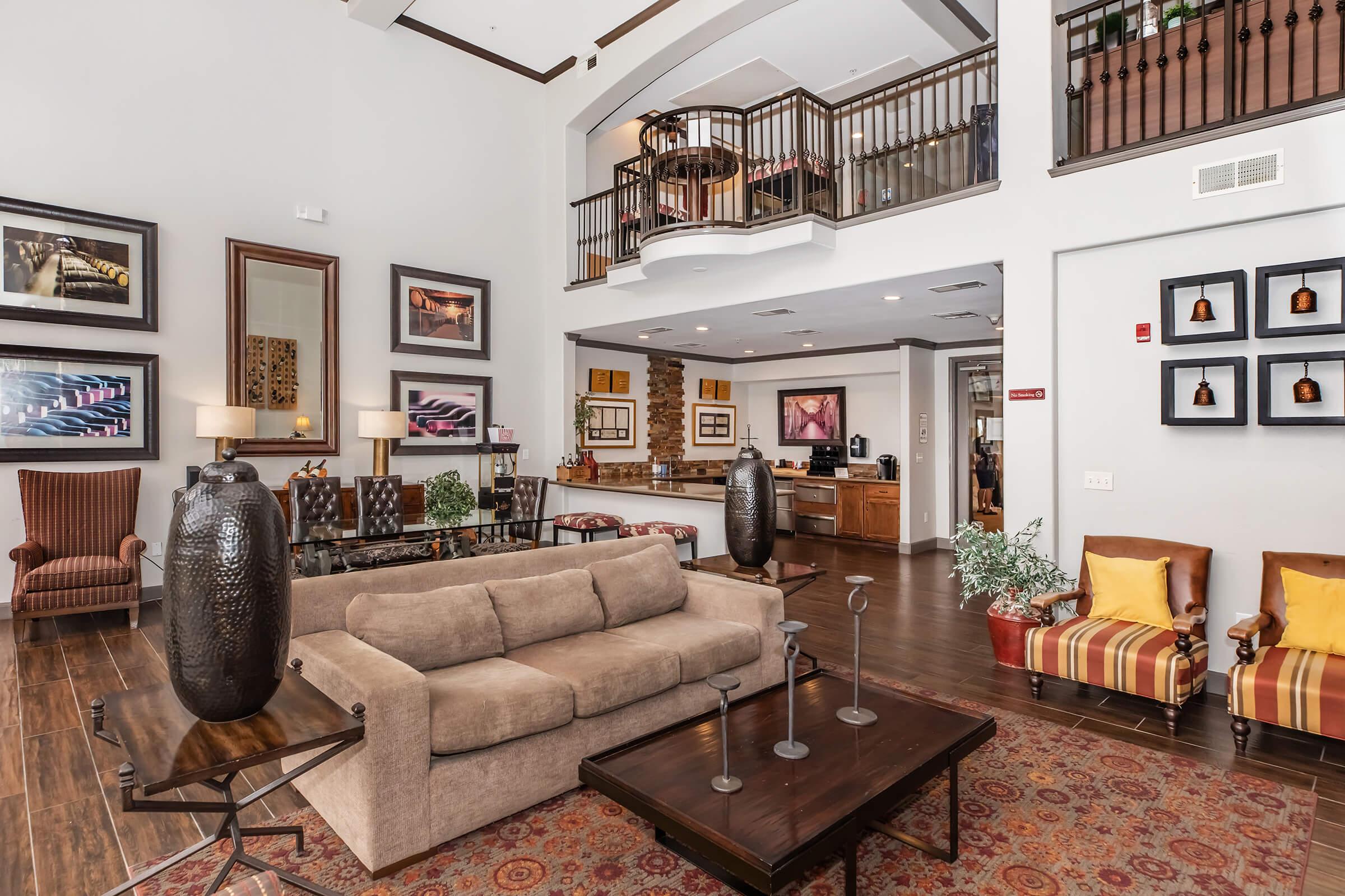Modern lobby area with high ceilings, featuring a beige sofa and decorative coffee table. The walls are adorned with framed artwork, and there are tall vases and indoor plants. A reception desk is visible in the background, along with a staircase leading to the upper level. Stools in a striped pattern add a pop of color.