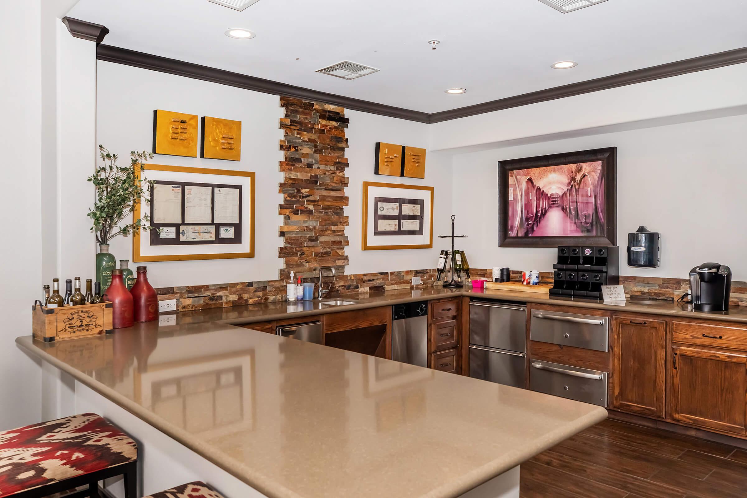 A modern kitchen area featuring a large countertop with bar stools, stainless steel appliances, and wooden cabinets. Decorative elements include framed artwork on the walls, stone accents, and various bottles displayed on the counter. The space is well-lit and showcases a warm, inviting atmosphere.